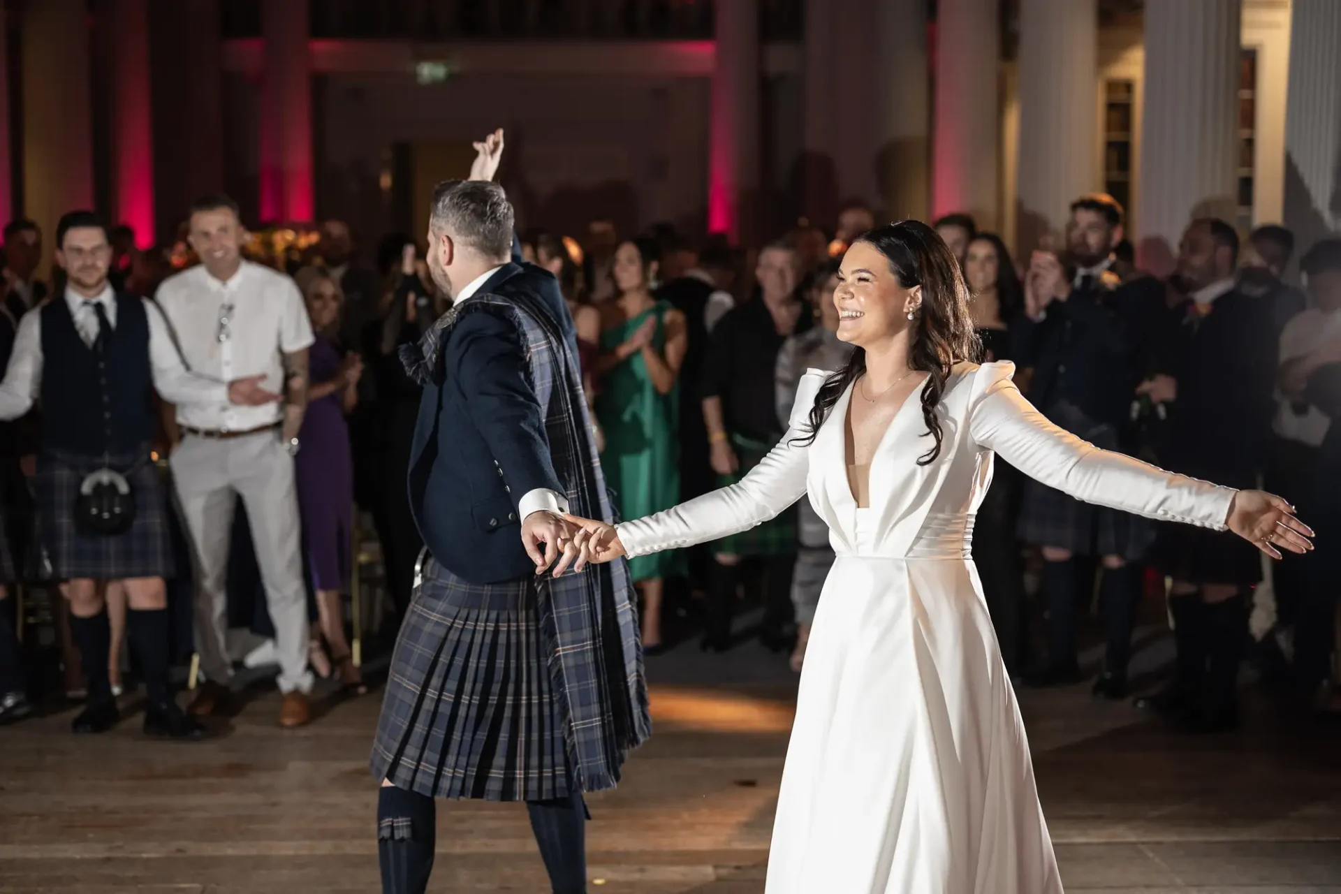 A bride and groom joyfully dance in front of guests at a wedding, with the groom wearing a kilt and the bride in a white dress.