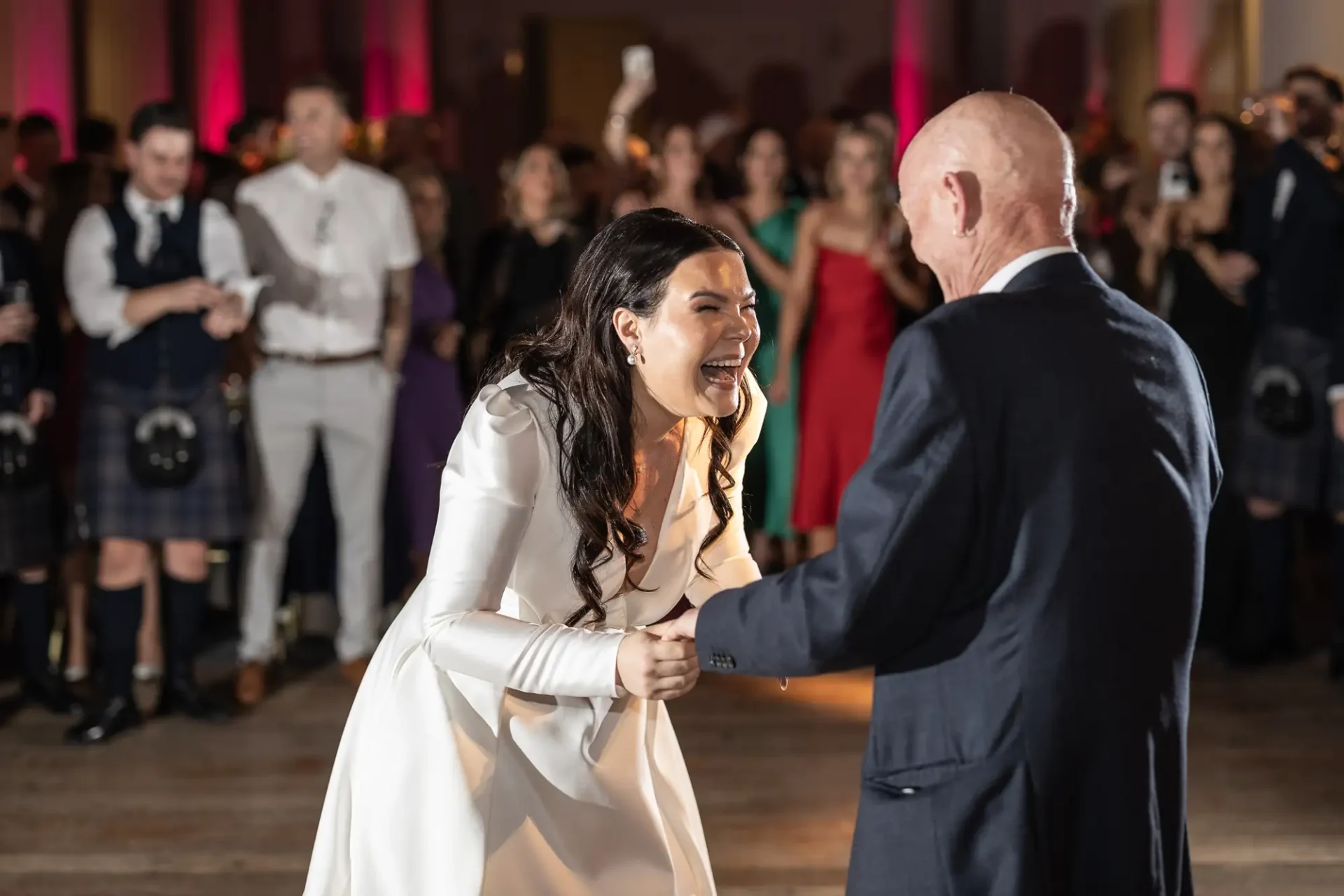 A bride in a white dress joyfully dances with a bald man in a suit at a wedding reception, surrounded by guests.
.