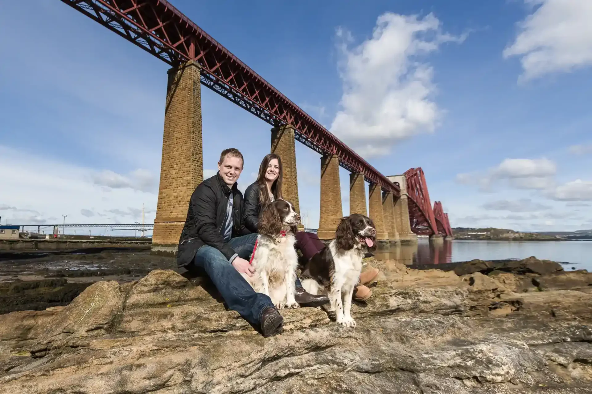 A man and woman sit on rocks with two dogs in front of a large railway bridge over water, under a blue sky with clouds.