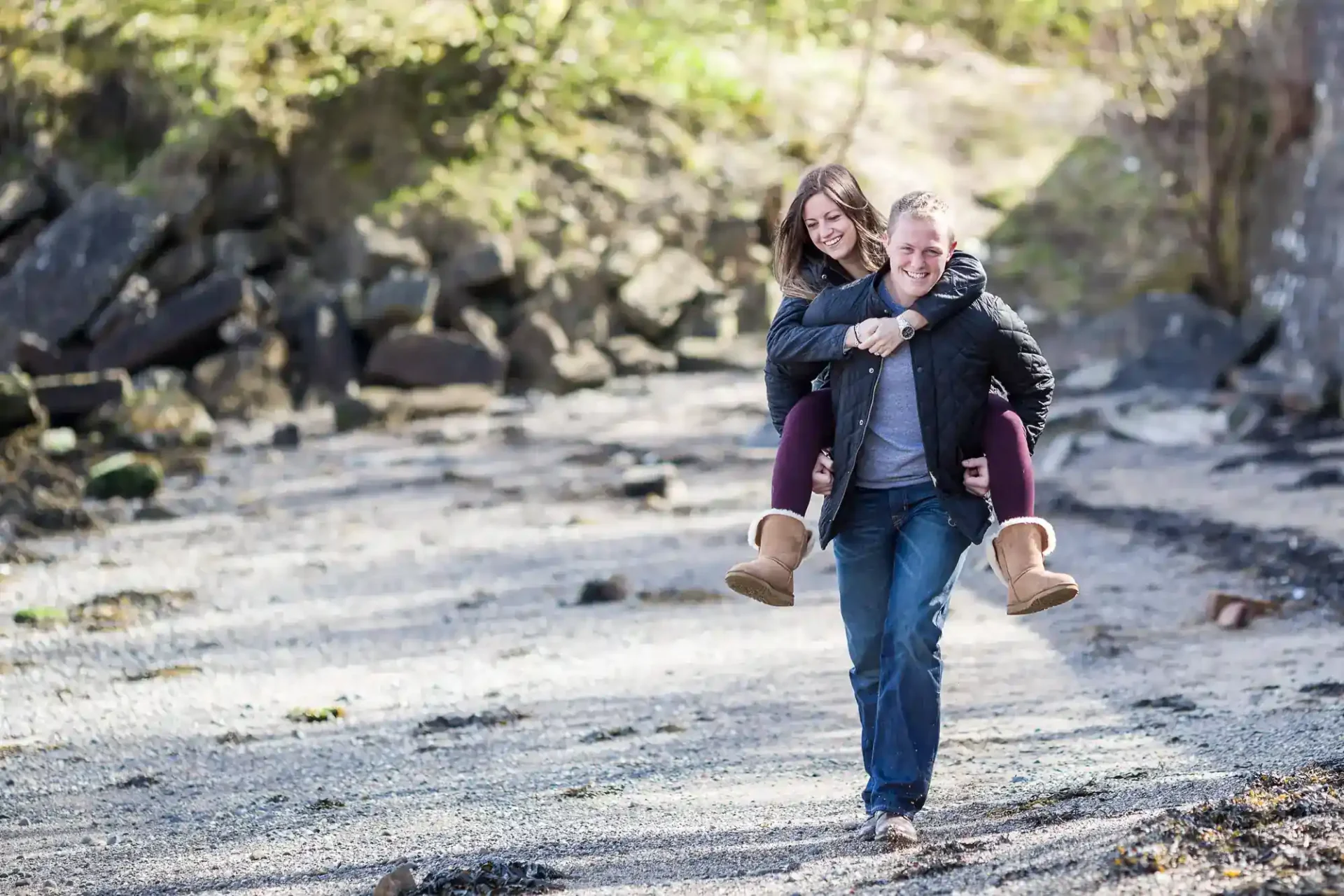 A person gives another person a piggyback ride on a rocky outdoor path, both smiling and dressed in casual clothes and boots. Trees and rocks are in the background.