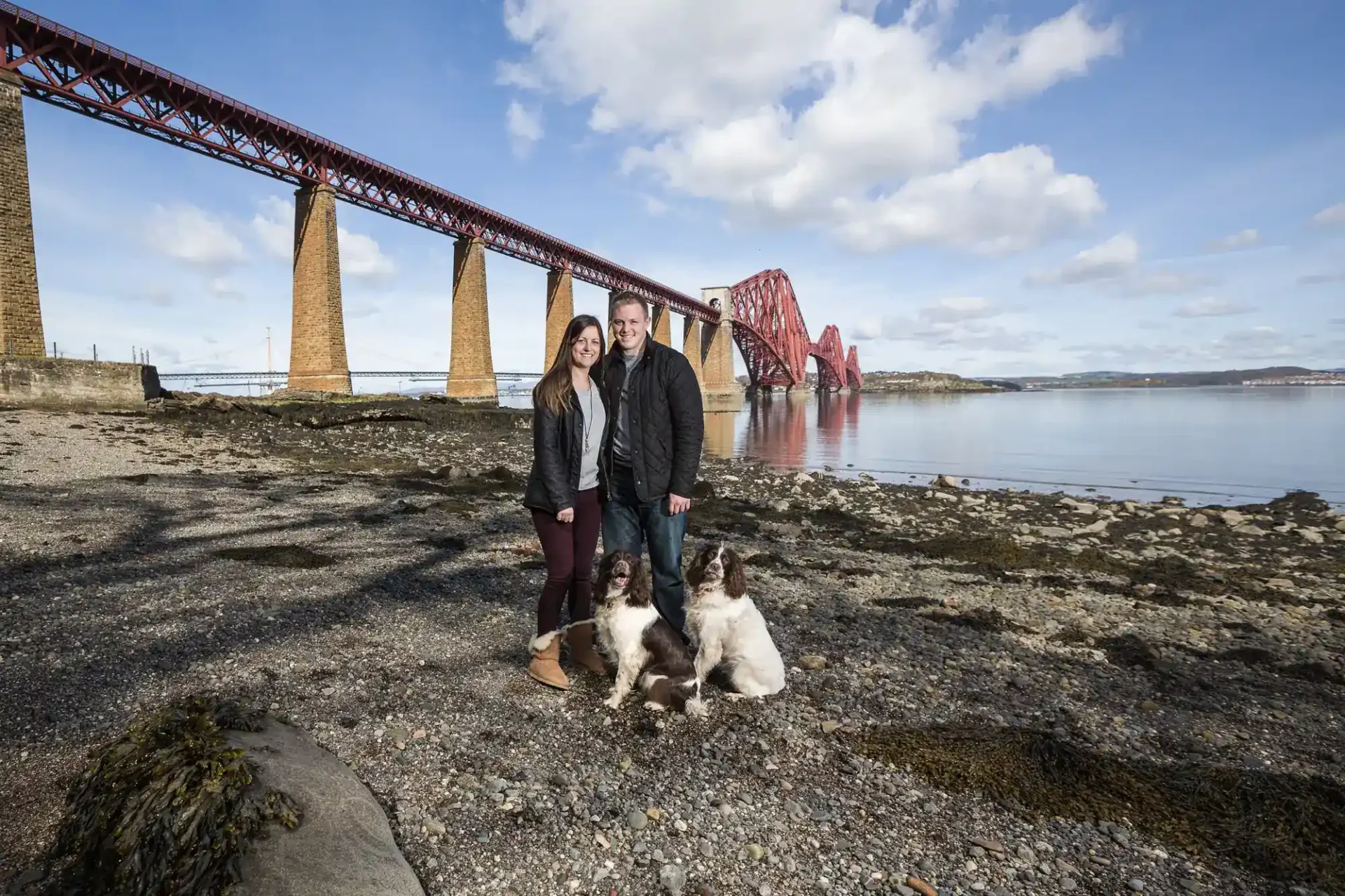 A couple stands on a rocky shore with two dogs, with a large red railway bridge spanning the water in the background under a partly cloudy sky.