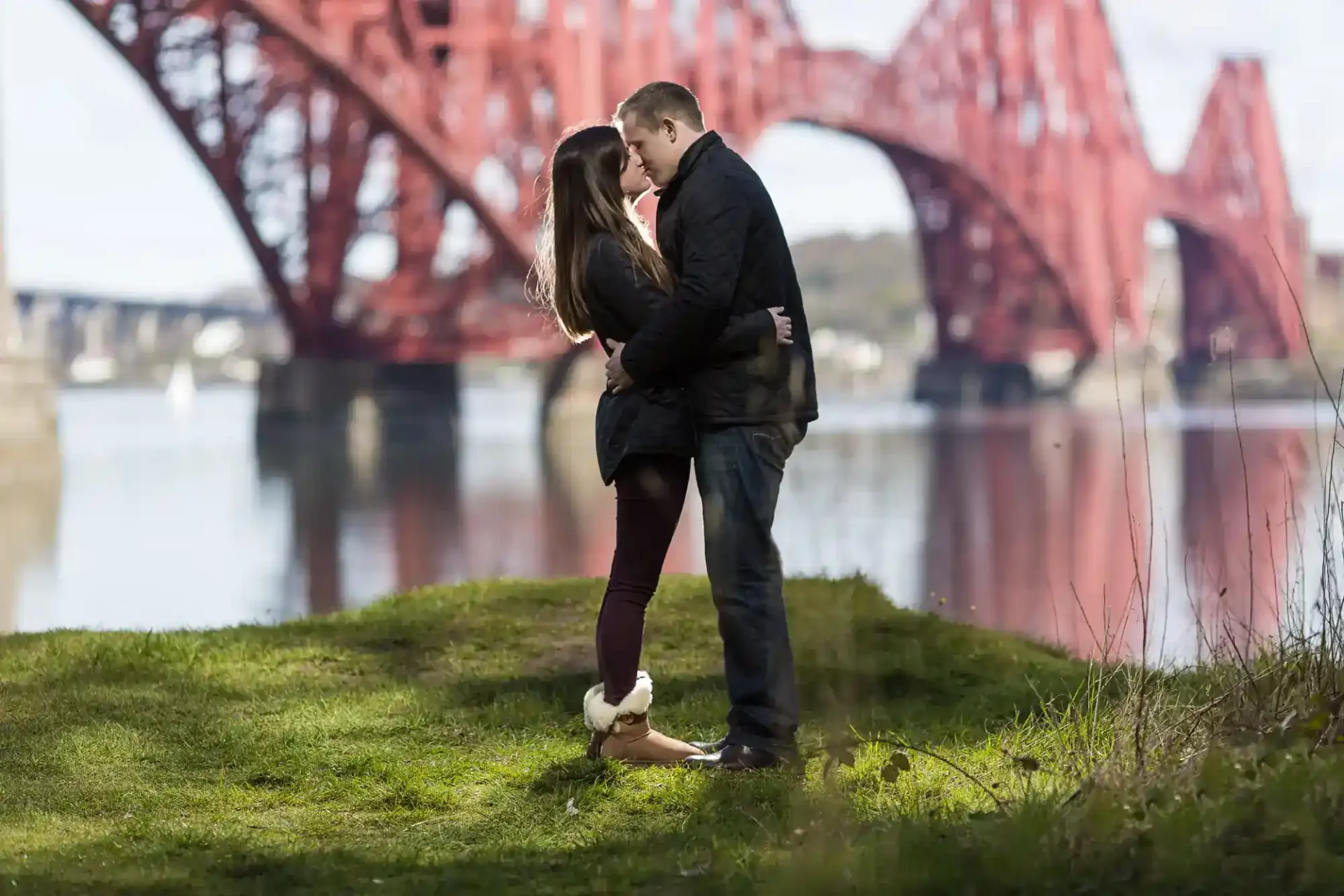 A couple stands closely together on grass by the water, with a large red steel bridge in the background.