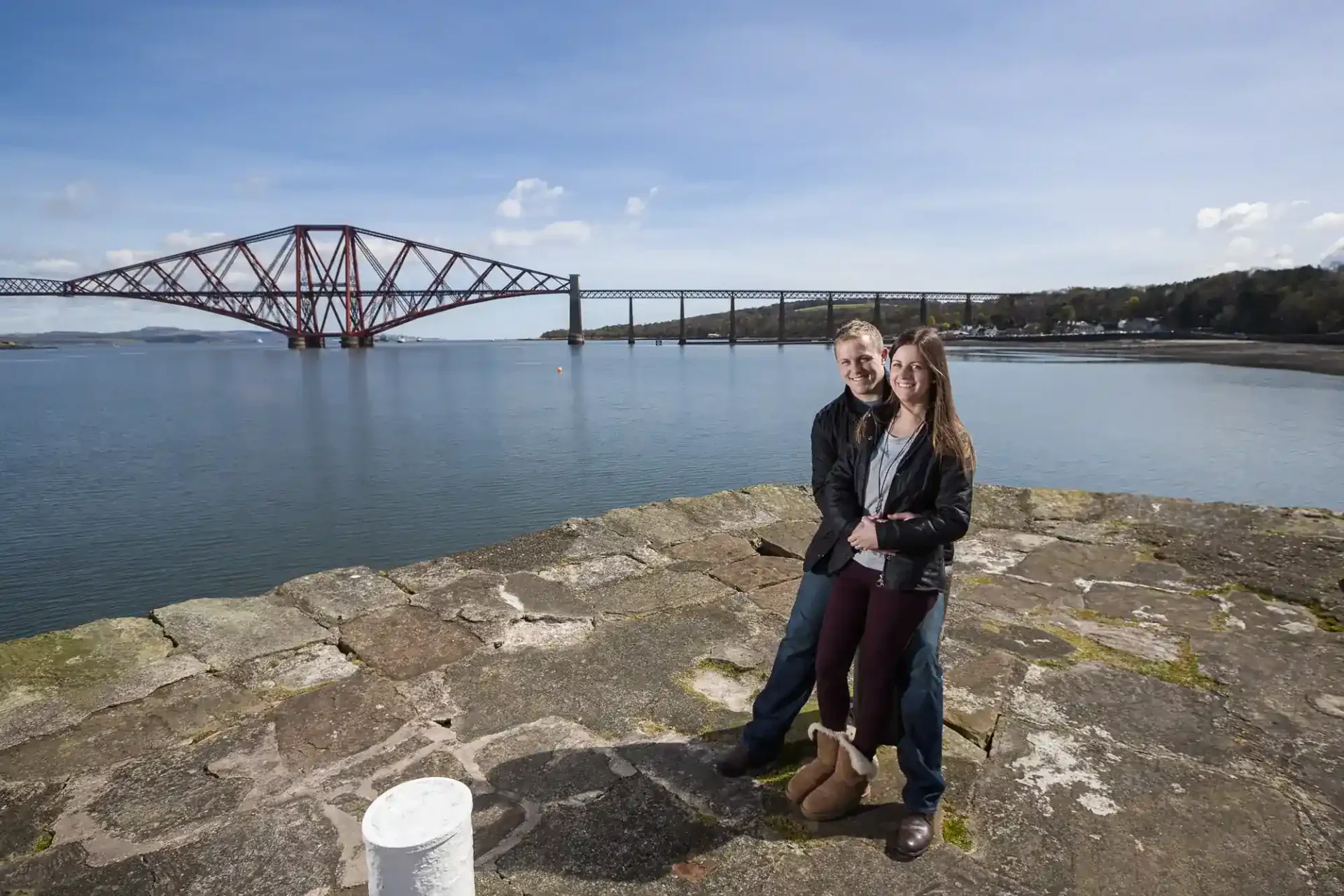 A smiling couple stands on a stone pier by the water, with the Forth Bridge visible in the background under a blue sky.