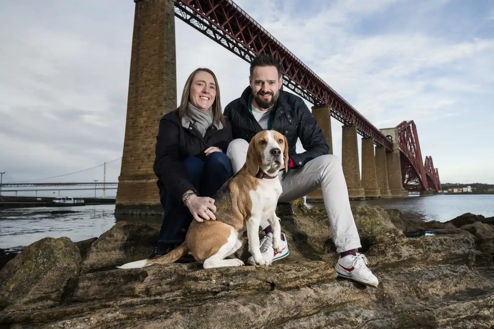 A woman and man sit on rocks with a beagle dog in front of a large red bridge over water, under a cloudy sky.