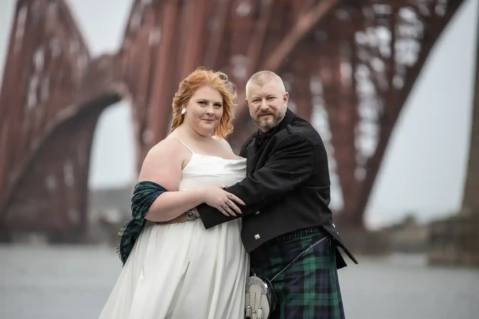 A couple poses together outdoors in formal attire, with the Forth Bridge in the background. The person on the right is wearing a kilt.