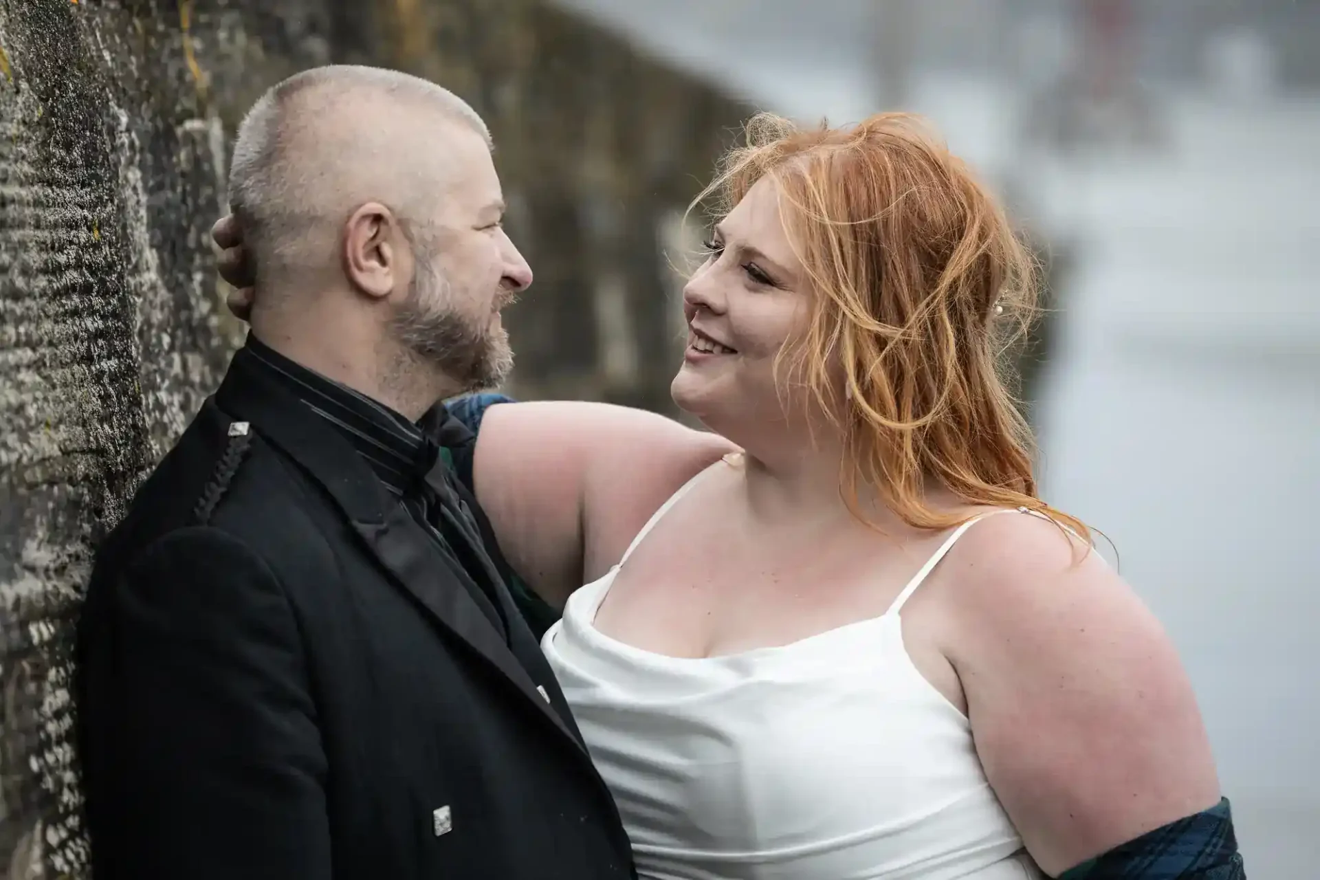 A couple stands close together by a stone wall outdoors; the woman in a white dress smiles at the man in a black suit.