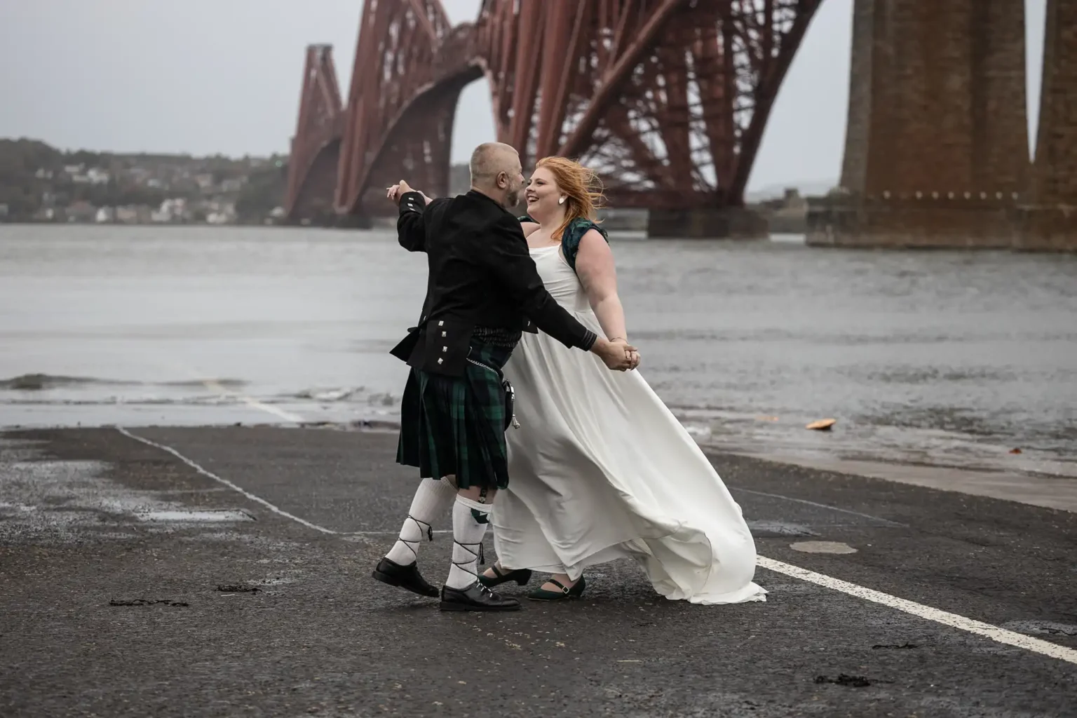 A couple, with the man in a kilt and the woman in a white dress, dance joyfully on a wet road near a large red bridge by the water.