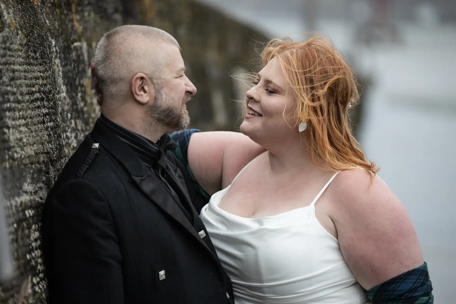A couple in formal attire, with the woman in a white dress and the man in a black suit, stand close together and smile at each other outdoors by a stone wall.