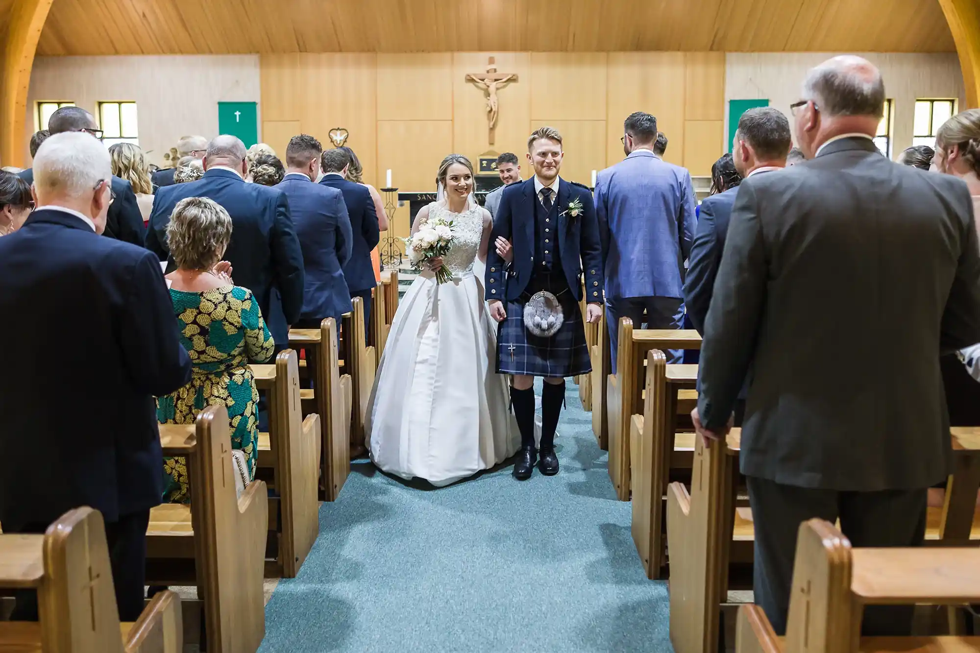 Bride in a white dress and groom in a kilt holding hands as they walk down the aisle of a church, surrounded by guests standing in the pews.