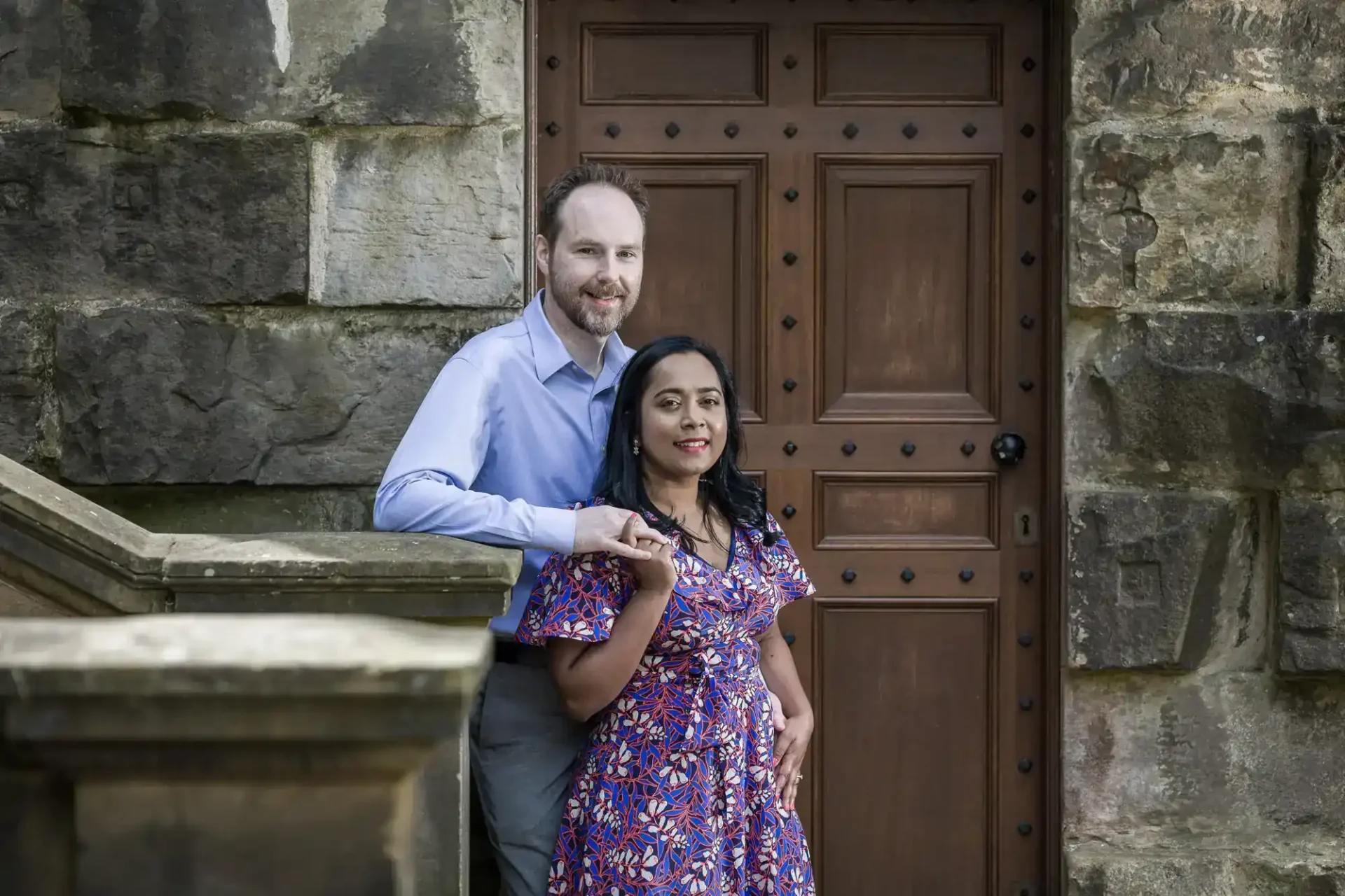 A man and woman pose together on stone steps in front of a wooden door and stone wall; the man leans casually while the woman stands in front.