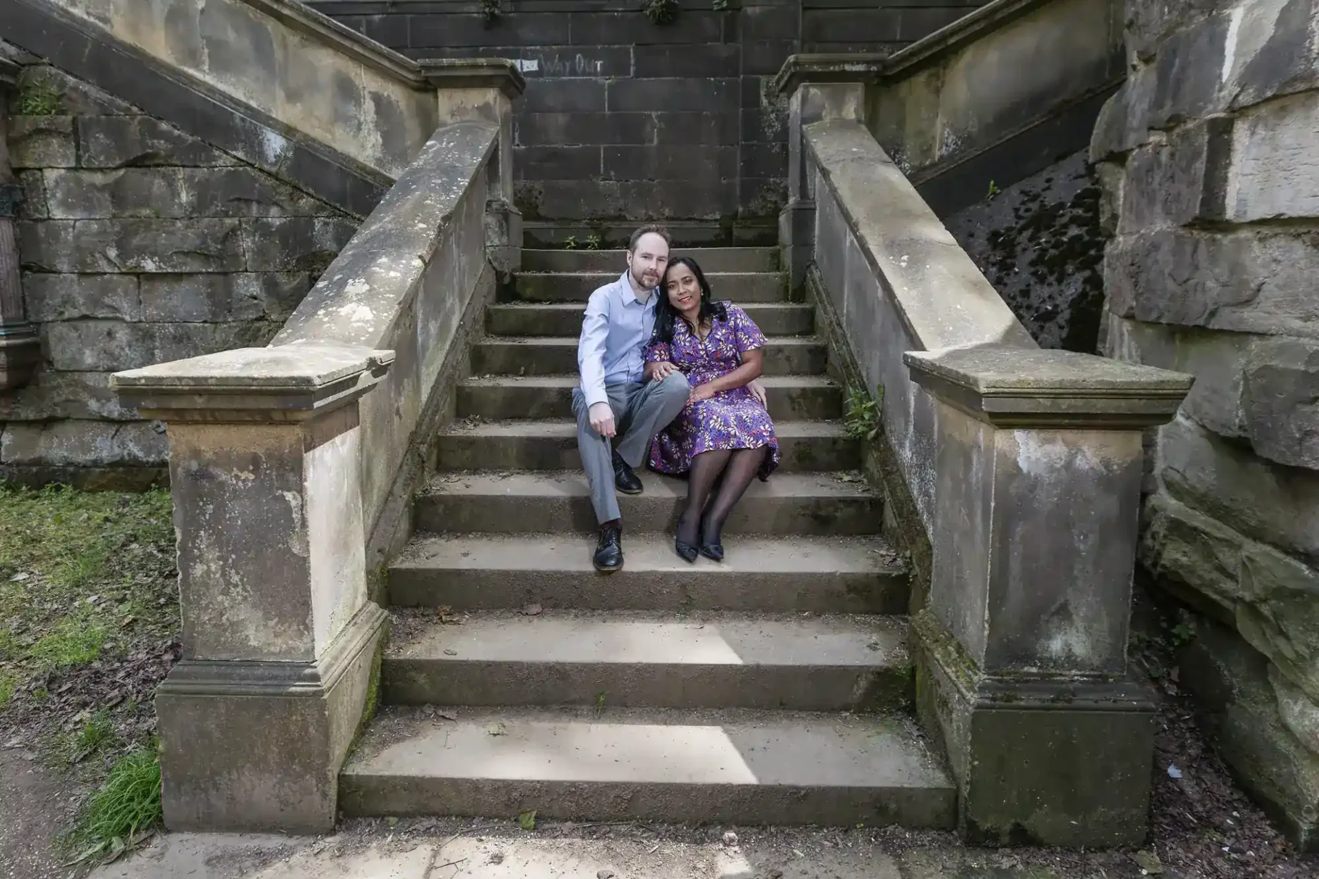 A man and woman sit together on outdoor stone steps, posing closely for a photo. The steps are bordered by stone railings and surrounded by weathered stone walls.