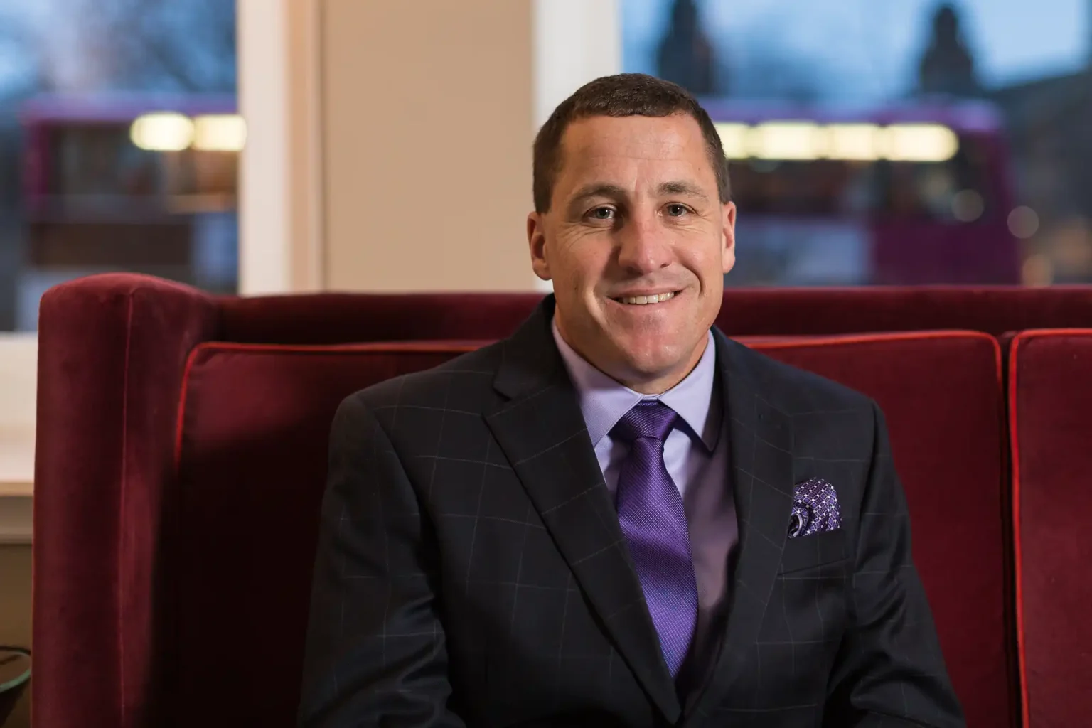 A man in a suit with a tie and pocket square smiles while sitting on a red sofa, with a window and blurred outdoor scene behind him.