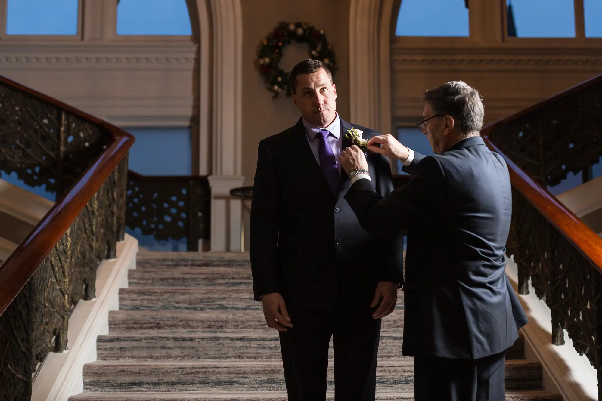 An older man fixes a boutonniere on a younger man's suit jacket in an elegant staircase hall adorned with a christmas wreath.