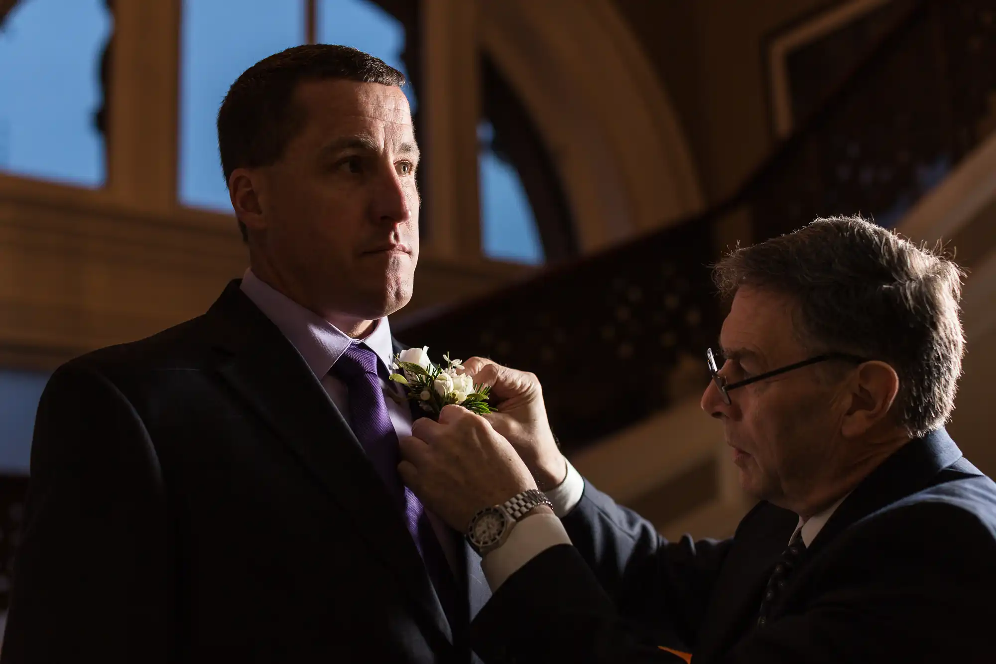 An older man adjusts a boutonniere on a younger man's suit jacket in a dimly lit room with wooden interiors.