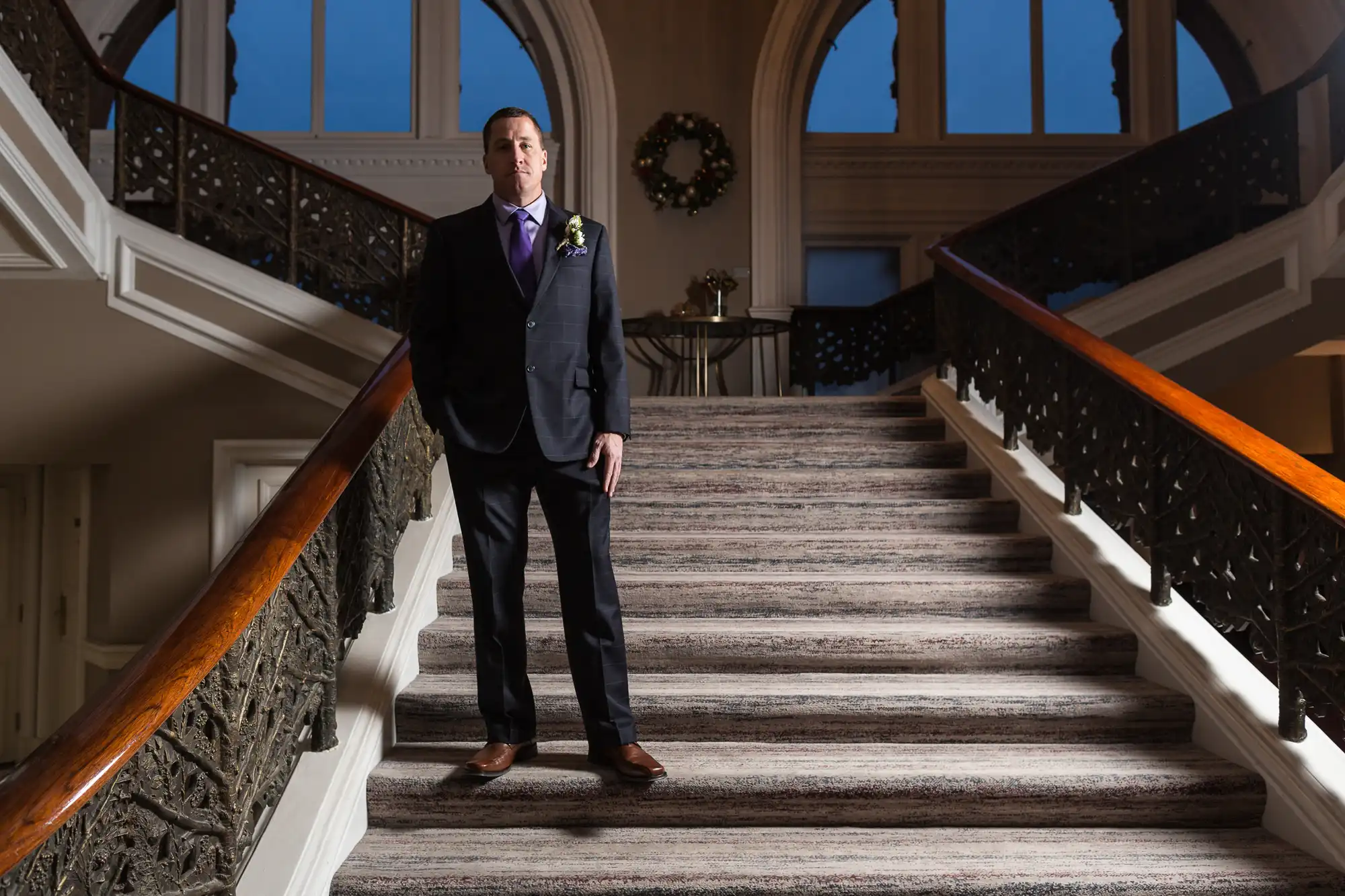 Man in a dark suit standing on a grand staircase with ornate railings inside an elegant building.