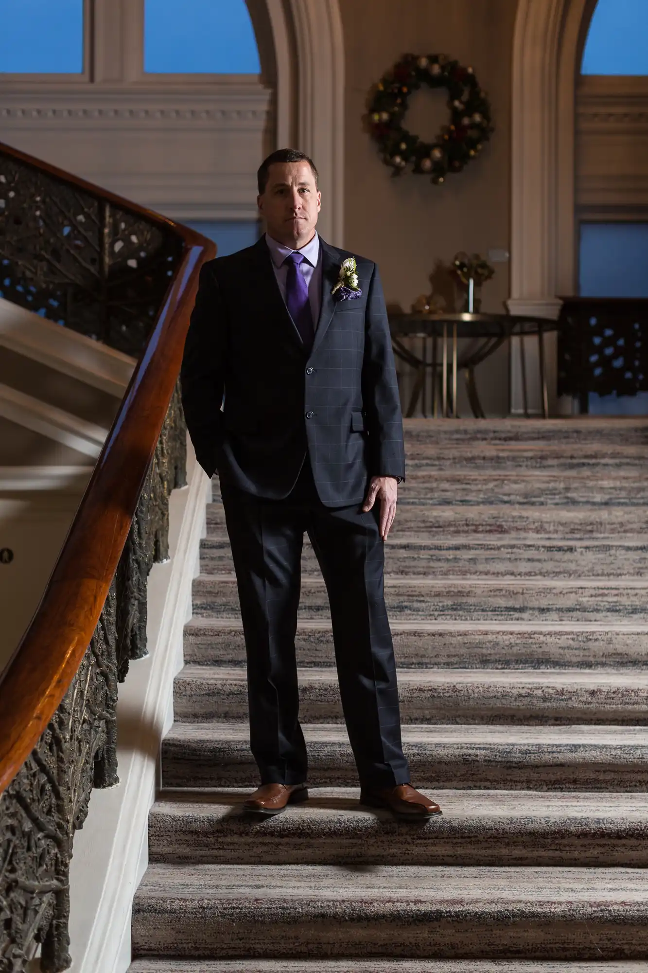 A man in a dark suit and purple tie stands solemnly on a grand staircase with a decorated wreath in the background.