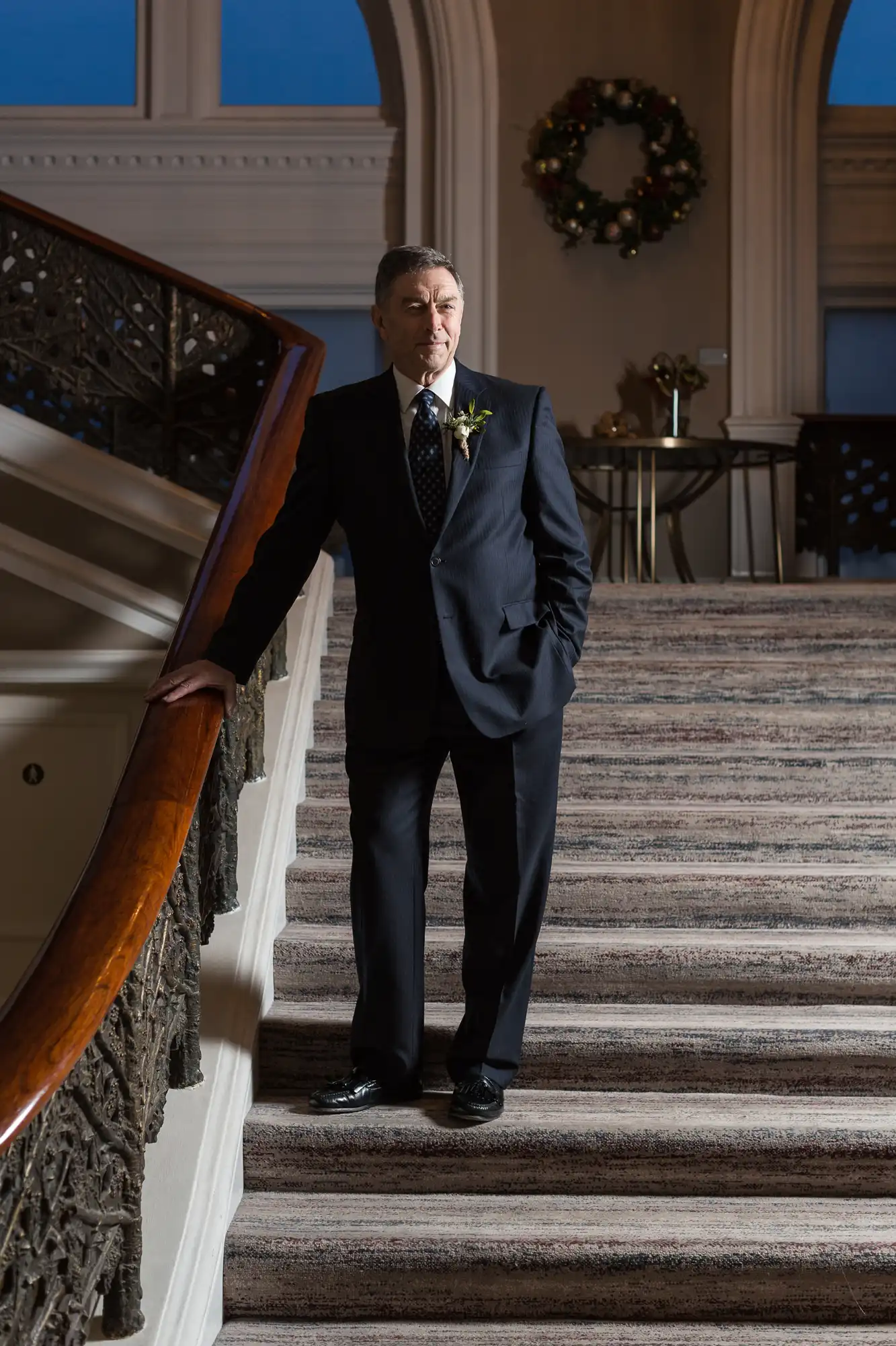 An older man in a dark suit and boutonniere stands on a grand staircase with a wooden railing, looking pensively to the side.