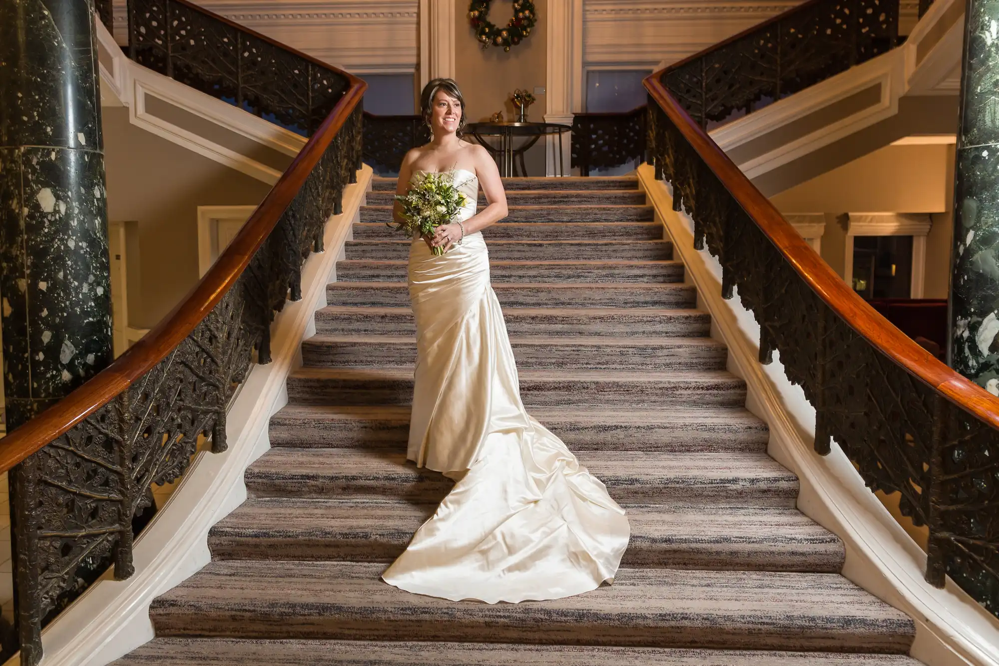 A bride in a strapless white gown holding a bouquet stands on an elegant staircase with dark bannisters and a carpeted runner, in a grand, opulent interior.
