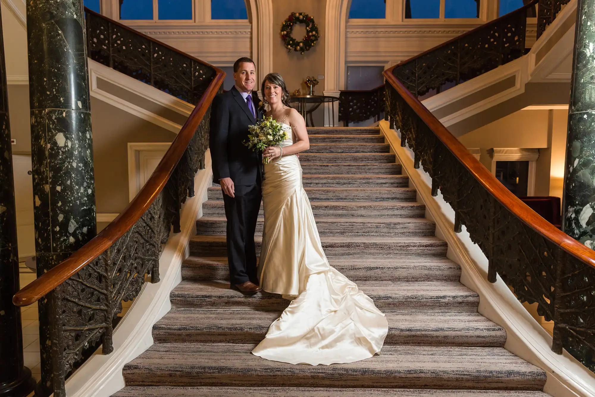 A bride and groom posing together on a grand staircase, the bride in a long cream dress and the groom in a dark suit.