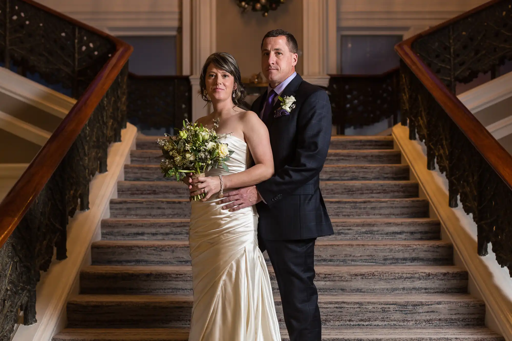 A bride and groom standing together on a staircase, the bride holding a bouquet, both looking slightly to the side.
