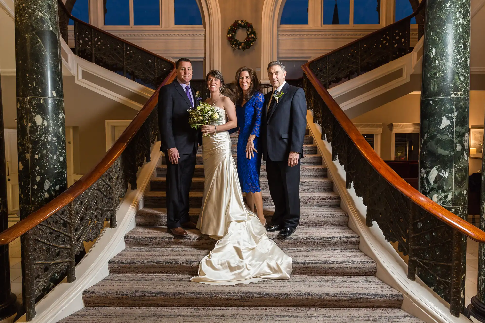 Two couples posing on an elegant staircase with marble railings inside a grand building, dressed in formal wedding attire.