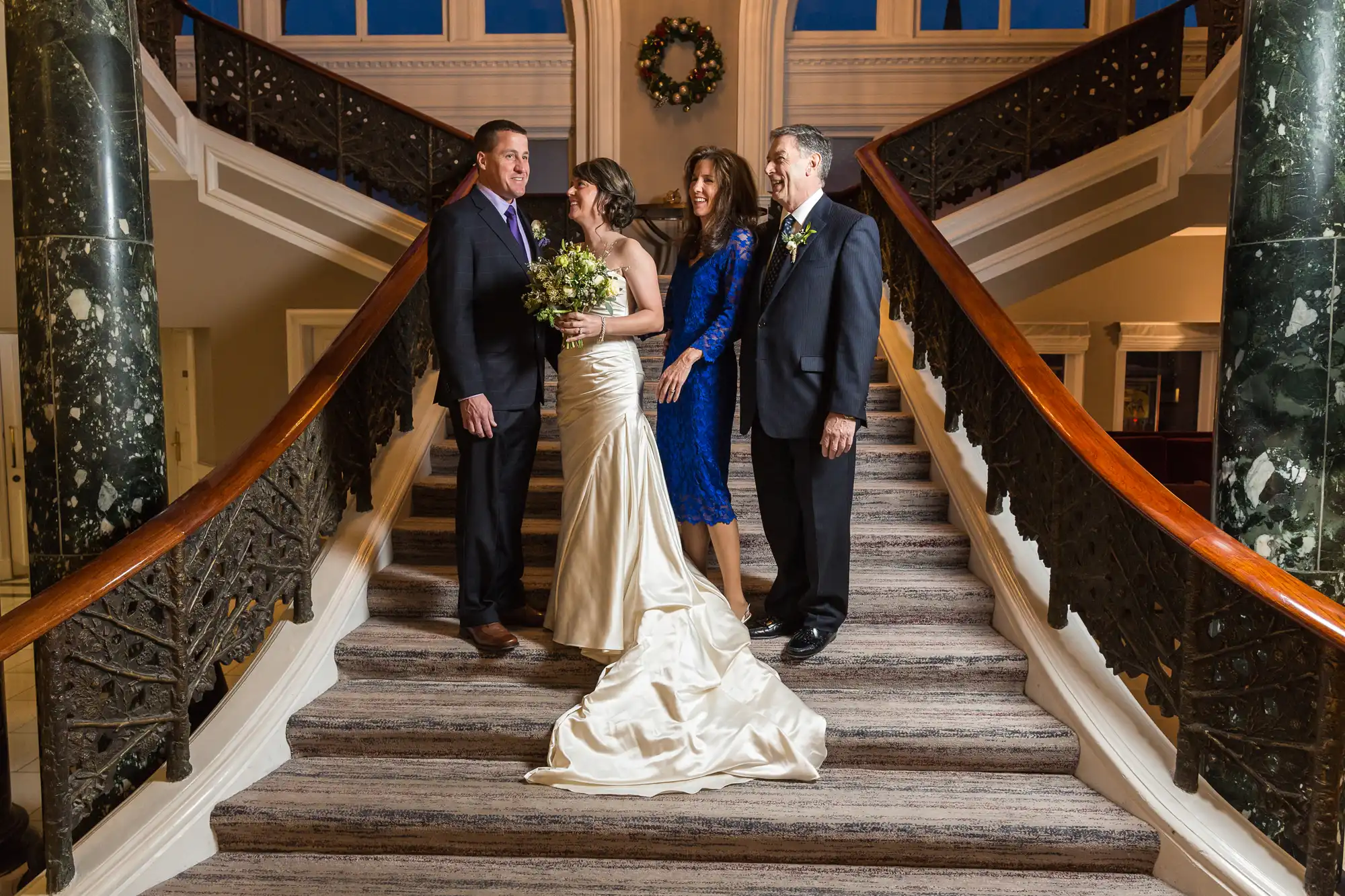A bride and groom descend a grand staircase with their parents, all smiling and dressed in formal attire.