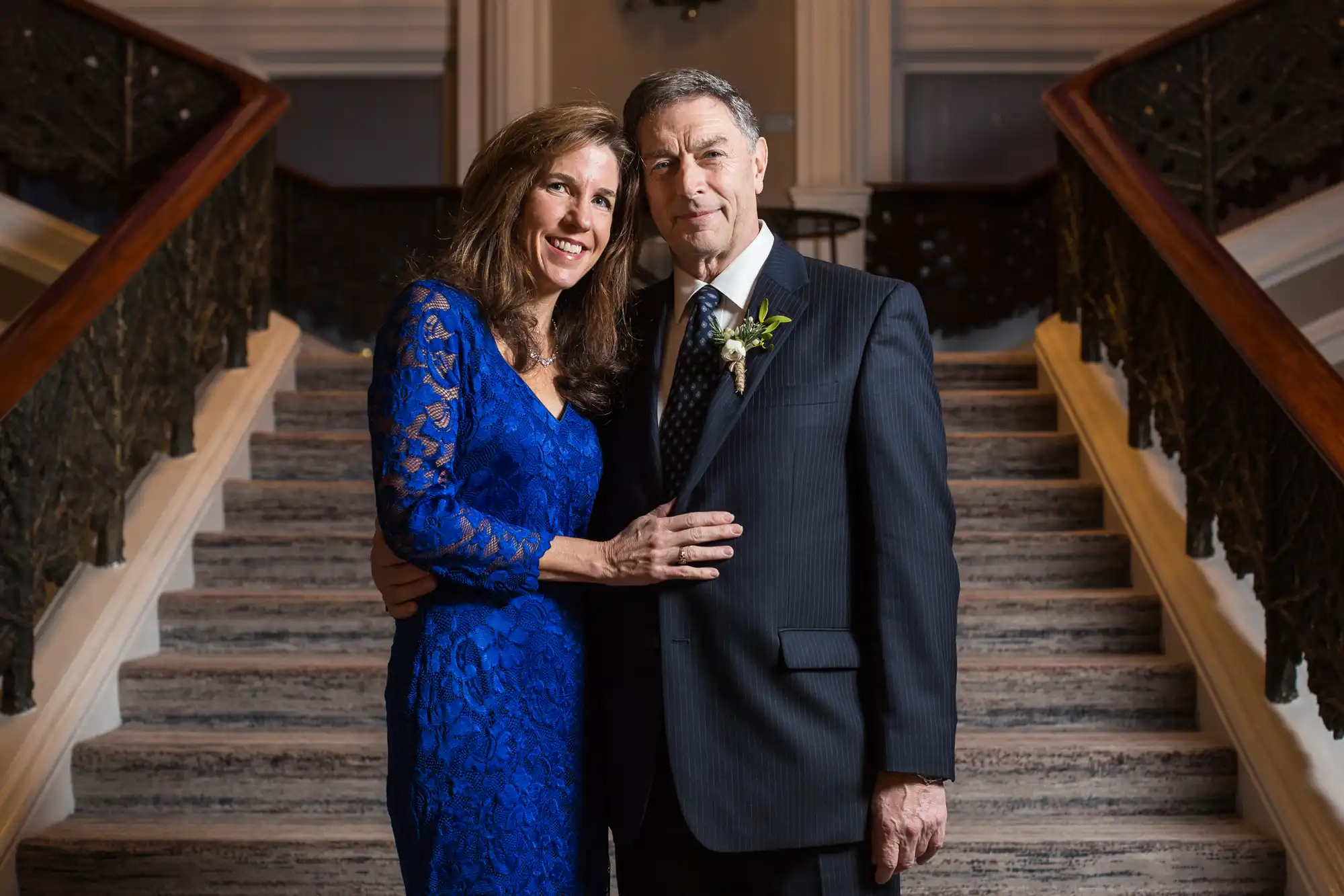 An elderly man in a suit and a woman in a blue dress smiling and posing together on a grand staircase.
