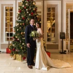 A couple in wedding attire posing in front of a tall christmas tree decorated with red and gold ornaments in a lobby.