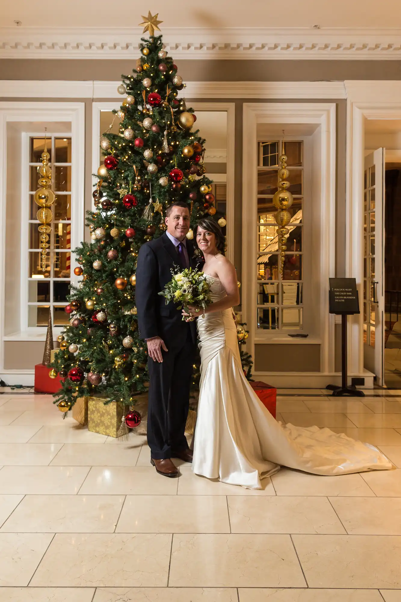A couple in wedding attire posing in front of a tall christmas tree decorated with red and gold ornaments in a lobby.