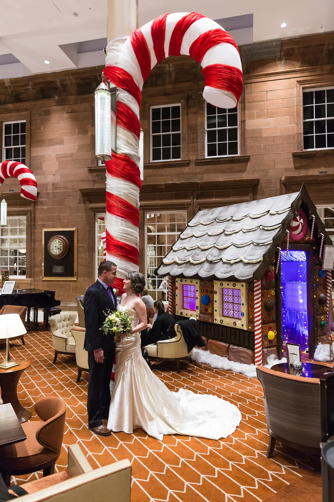 Newlywed couple posing in a hotel lobby with a large candy cane arch and a gingerbread house in the background.