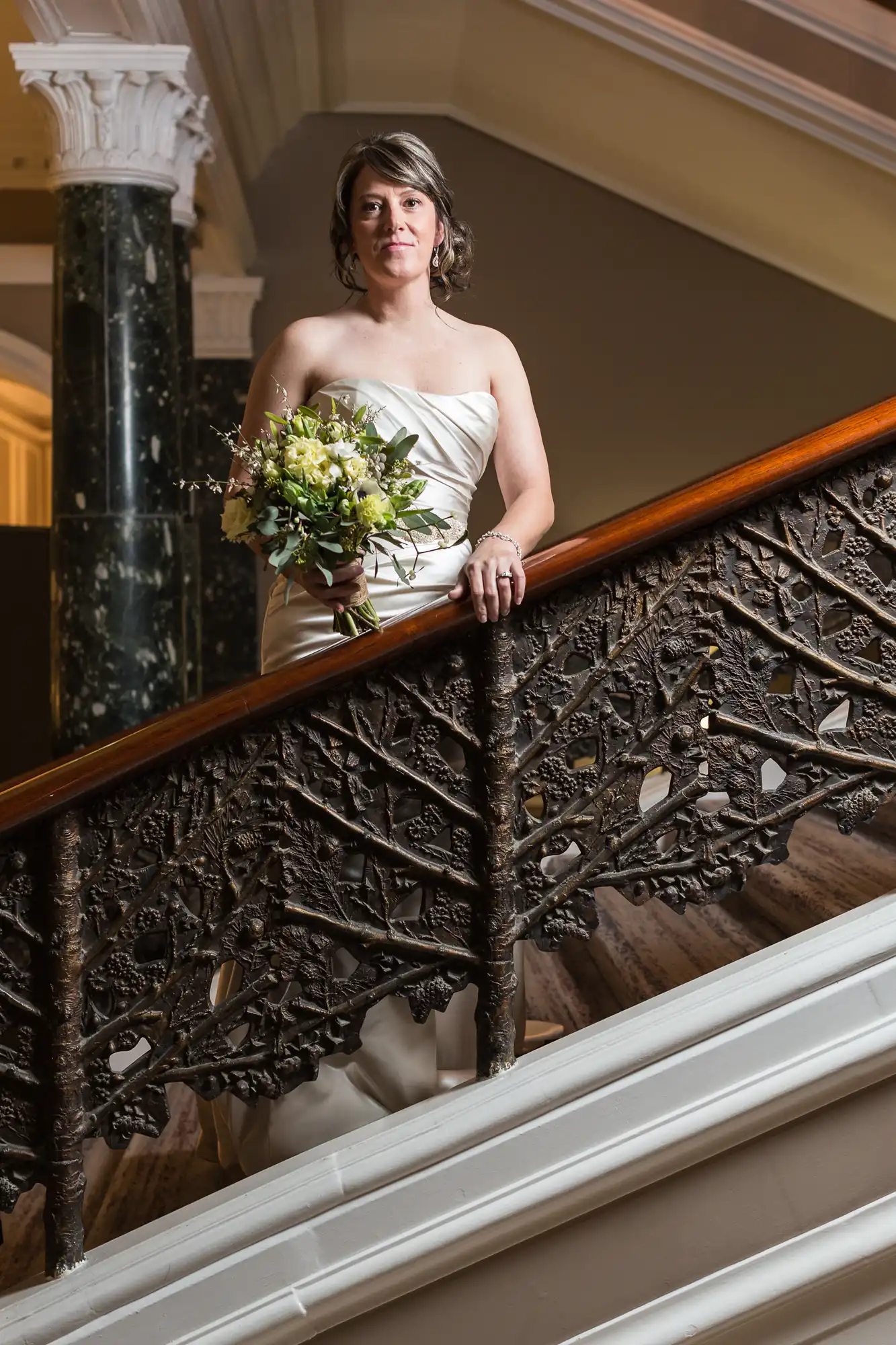 A bride in a strapless white dress, holding a bouquet, standing on a staircase with ornate ironwork.