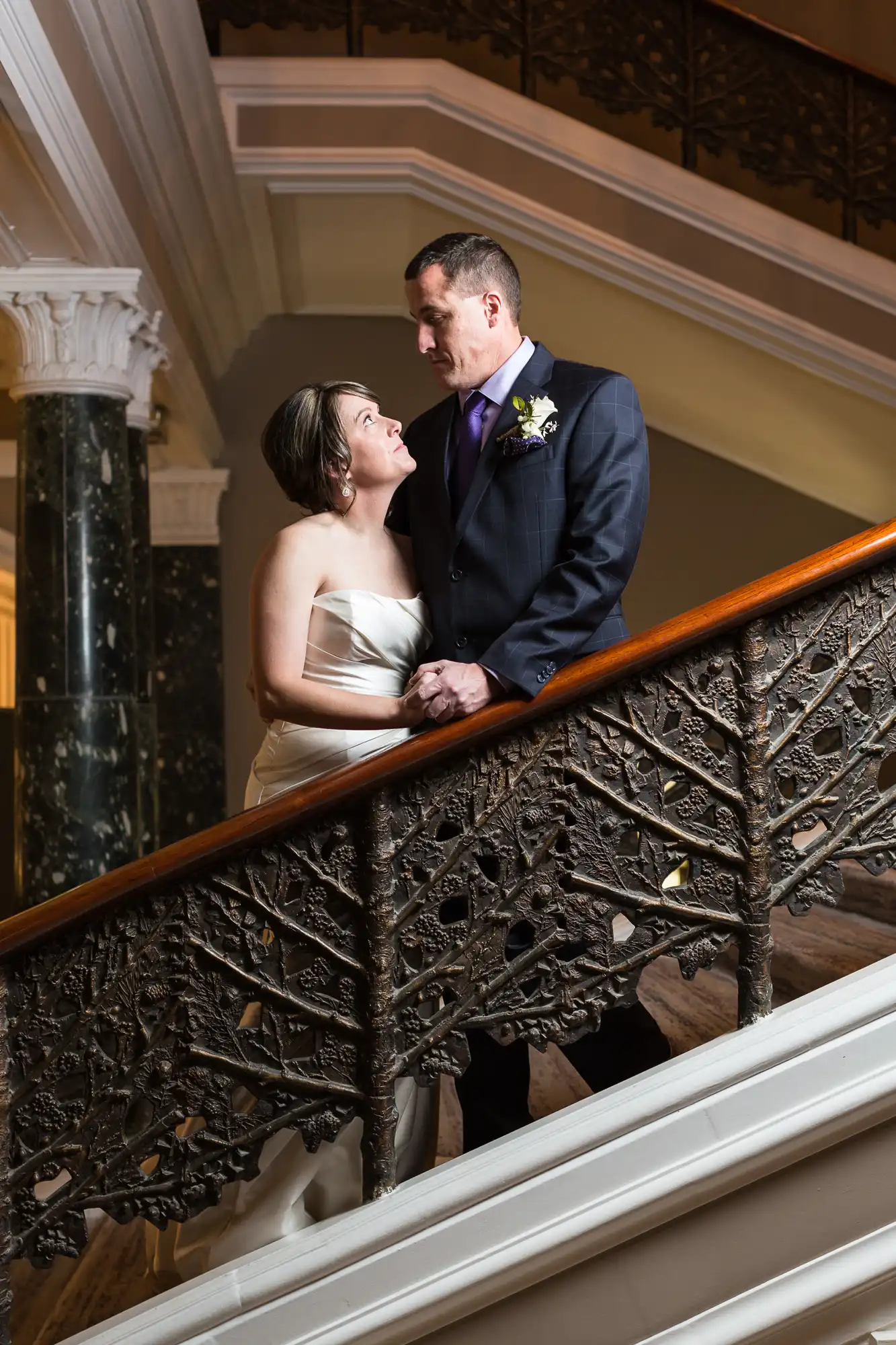 A couple in wedding attire affectionately gaze at each other while standing on an ornate staircase with intricate railings.