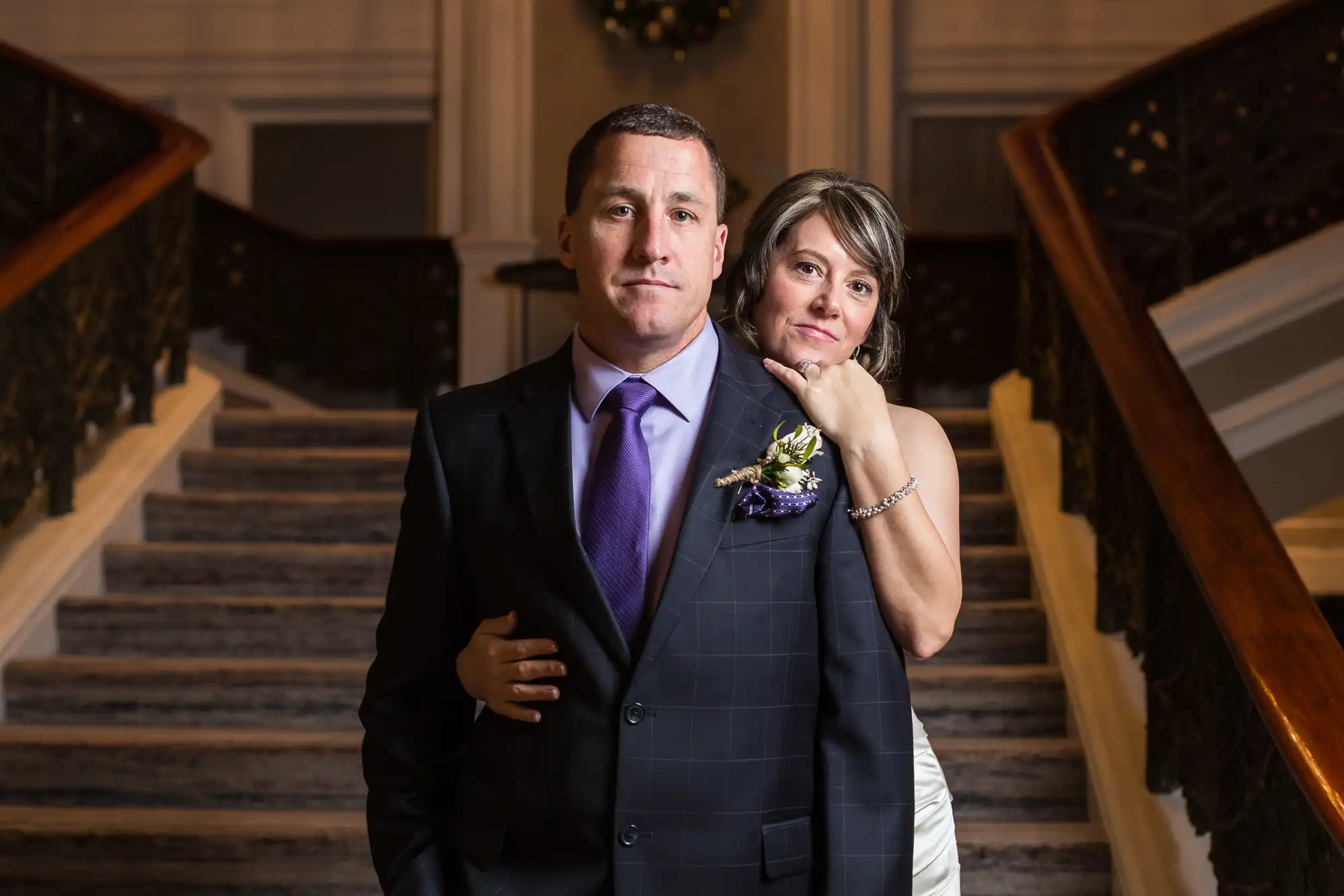 A couple dressed in formal attire stands in a grand hallway with staircases on either side, looking directly at the camera.