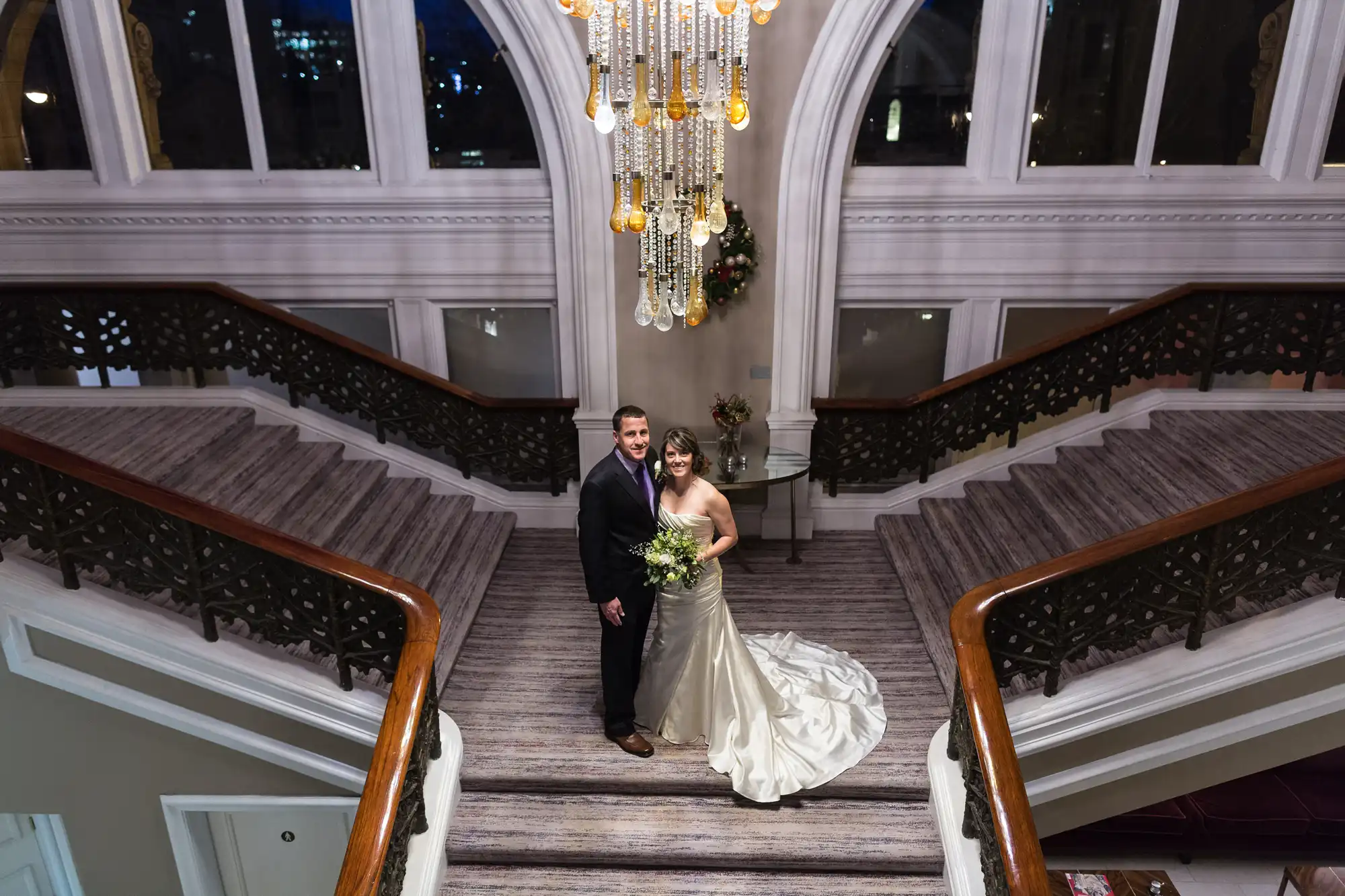 A newly married couple stands smiling on an ornate staircase under a chandelier, in an elegant building.