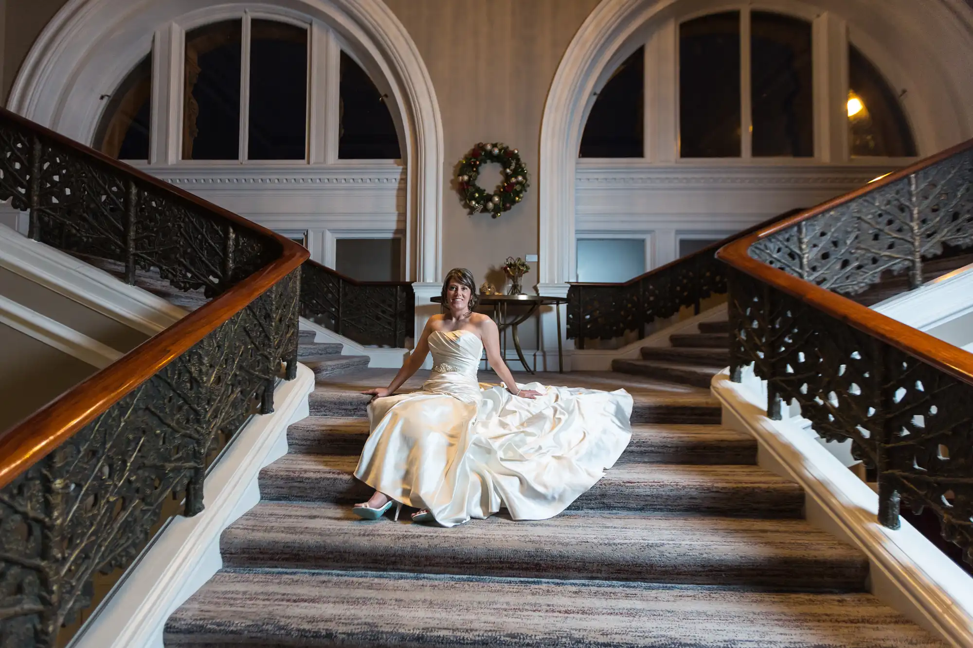 A woman in a white dress sits on a carpeted staircase between ornate metal railings, with large windows and a wreath in the background.
