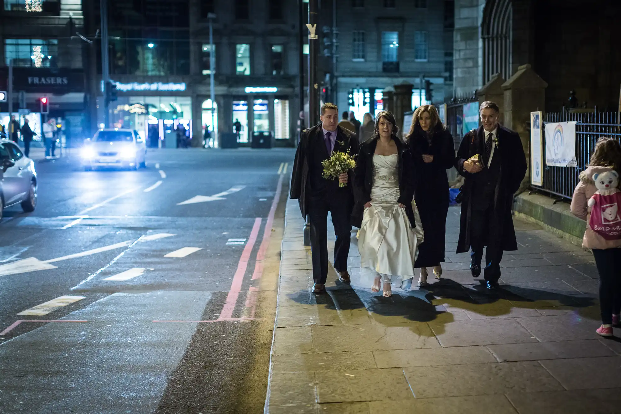 A wedding party walking along a city street at night, with a bride in a white dress, flanked by two men in suits and a woman in black, nearby a lit storefront.