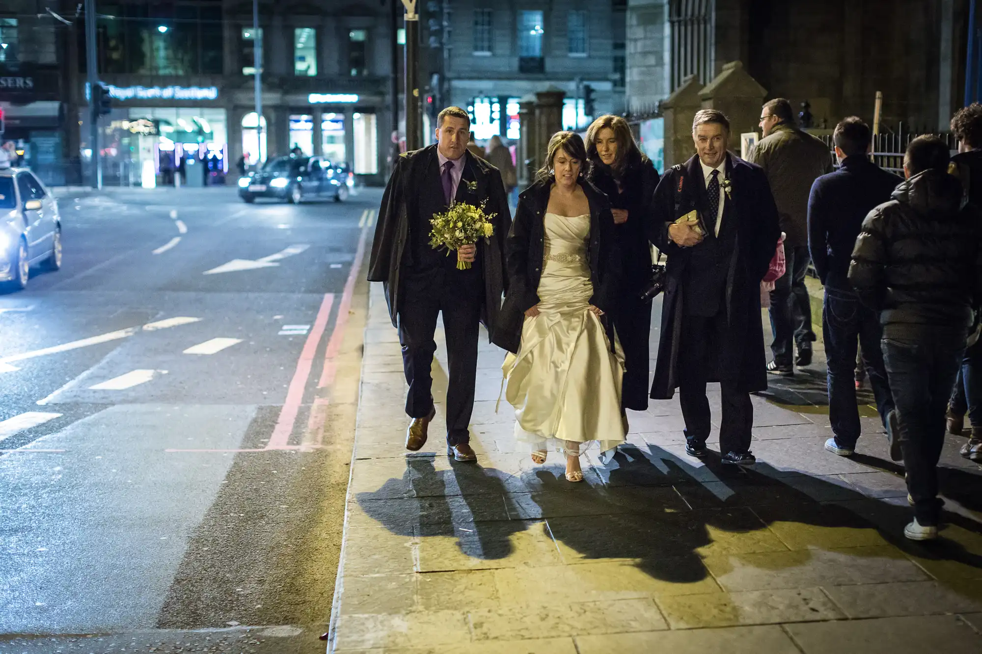 A newlywed couple with two companions crosses a city street at night, with bustling traffic and street lights in the background.