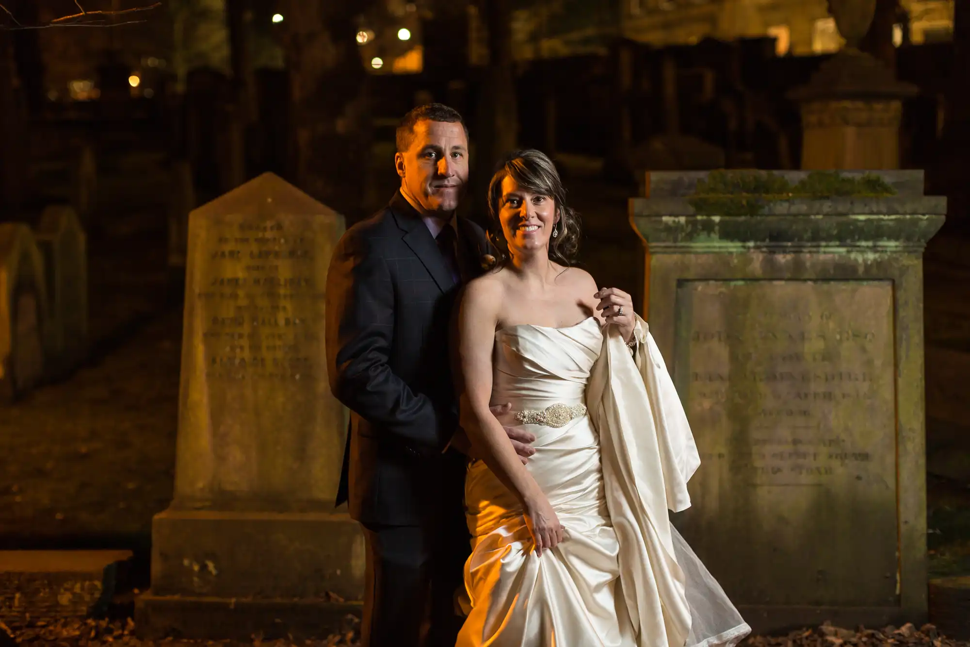 A bride and groom posing at night in a cemetery, standing between tombstones, the woman in a white dress and the man in a dark suit.