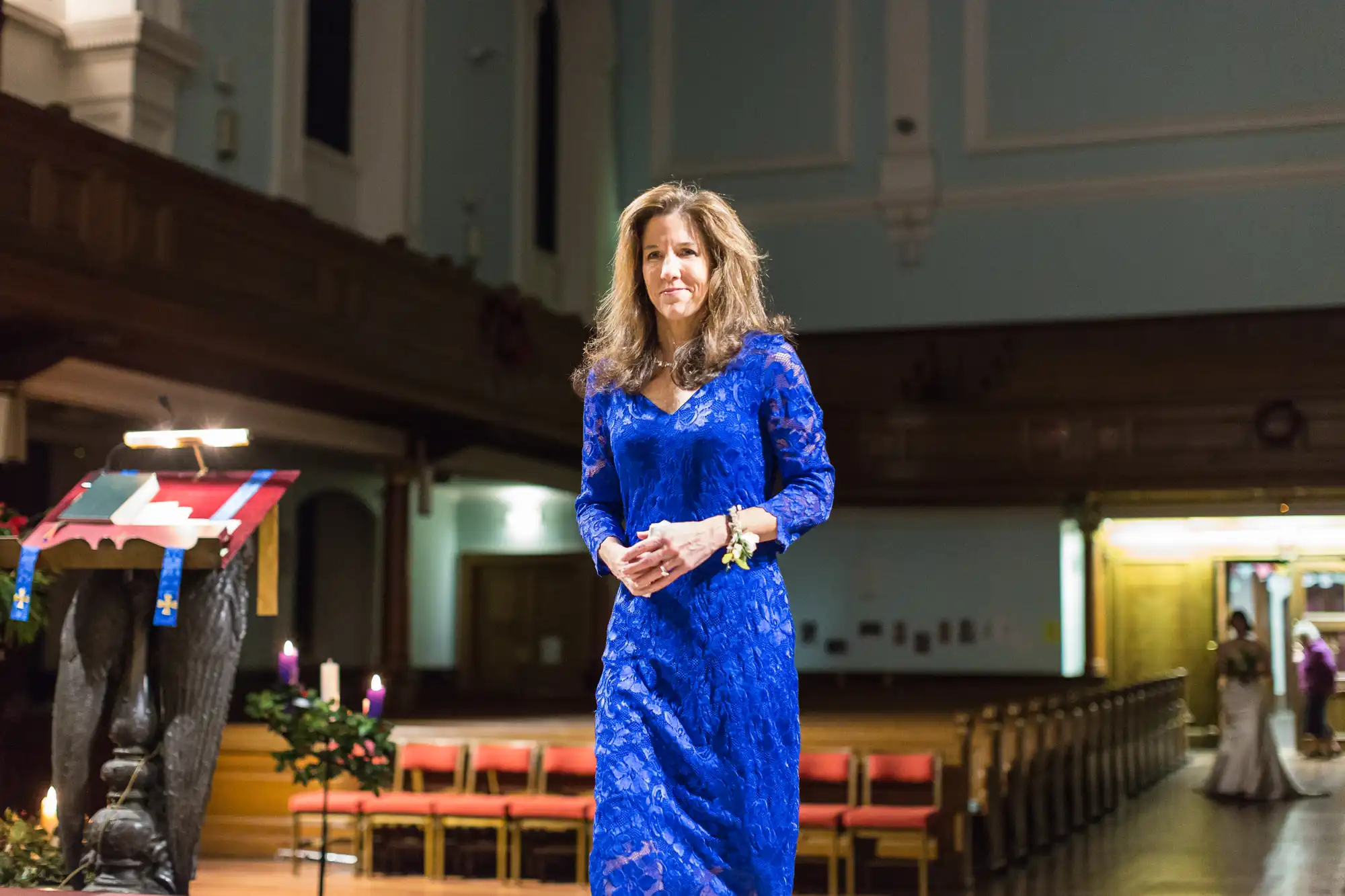 A woman in a blue lace dress stands in the aisle of a church, with pews and soft lighting in the background.