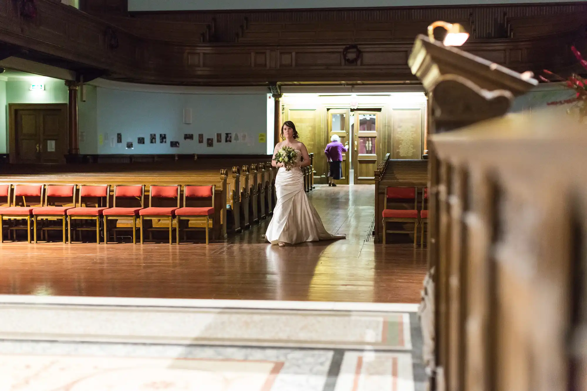 A bride in a white gown holding a bouquet walks down the aisle of an old hall with wooden chairs lined up along the sides.