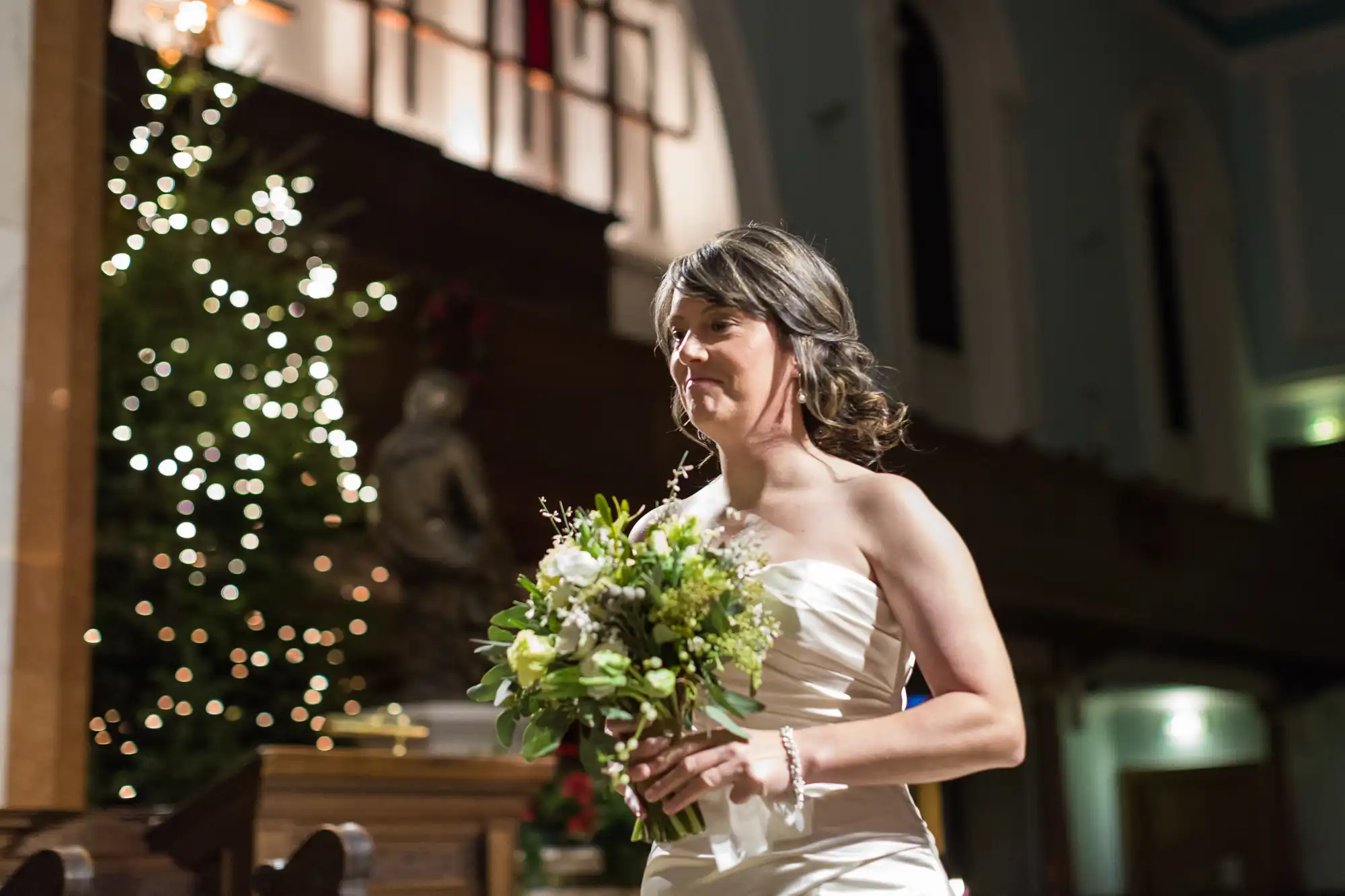 A bride in a white dress holding a bouquet walks down the aisle in a church decorated with string lights.