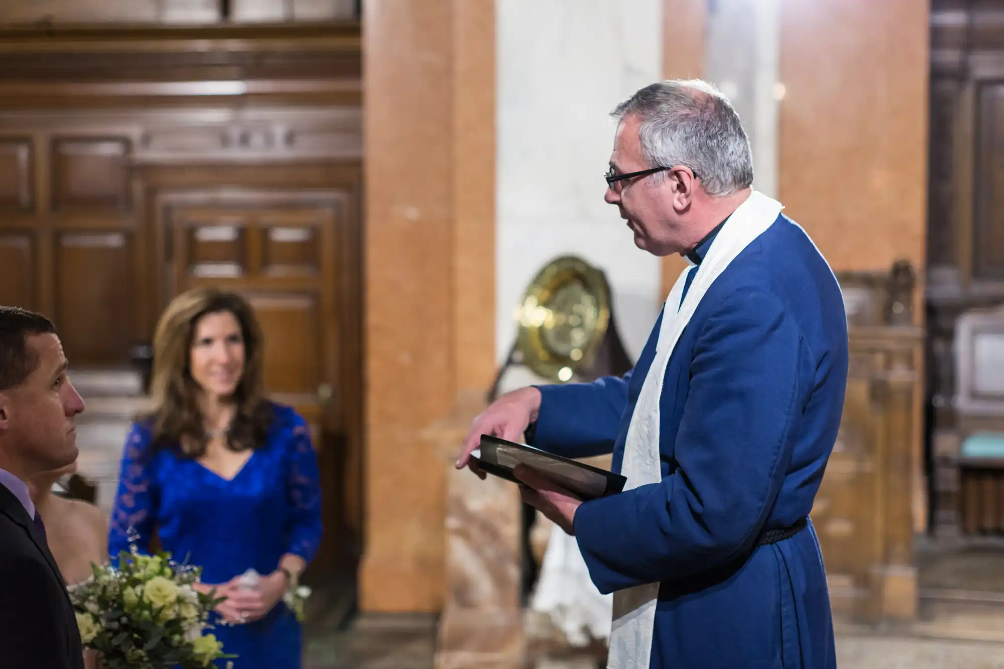 An older man in formal attire holding a book speaks to a couple in front of him inside a church, with a woman in a blue dress looking on.