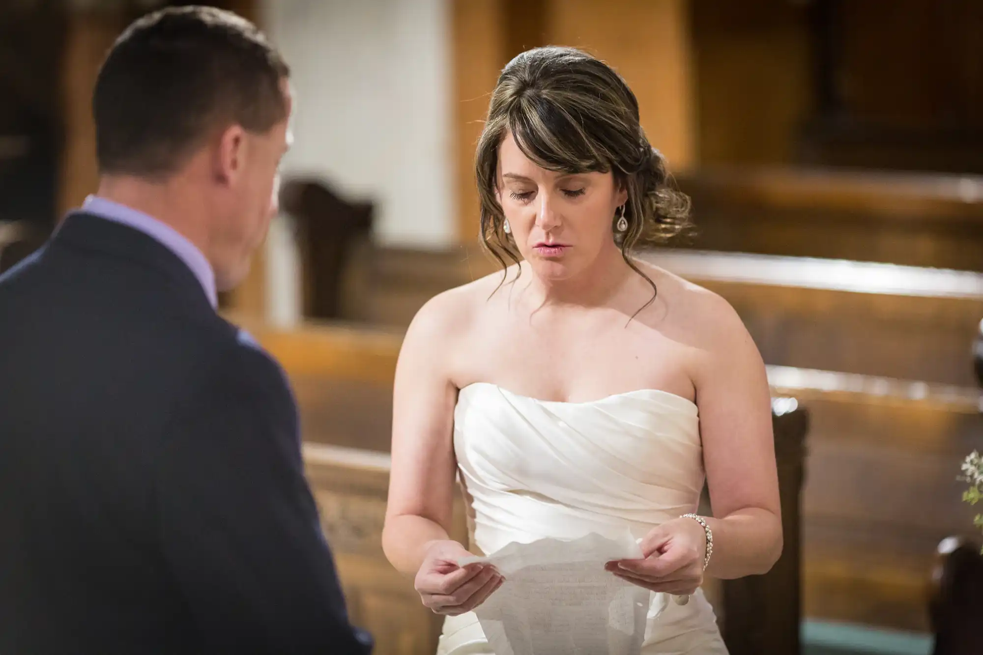 A bride in a white strapless gown reads her vows to the groom inside a church, holding a paper, with expressions of emotion on her face.