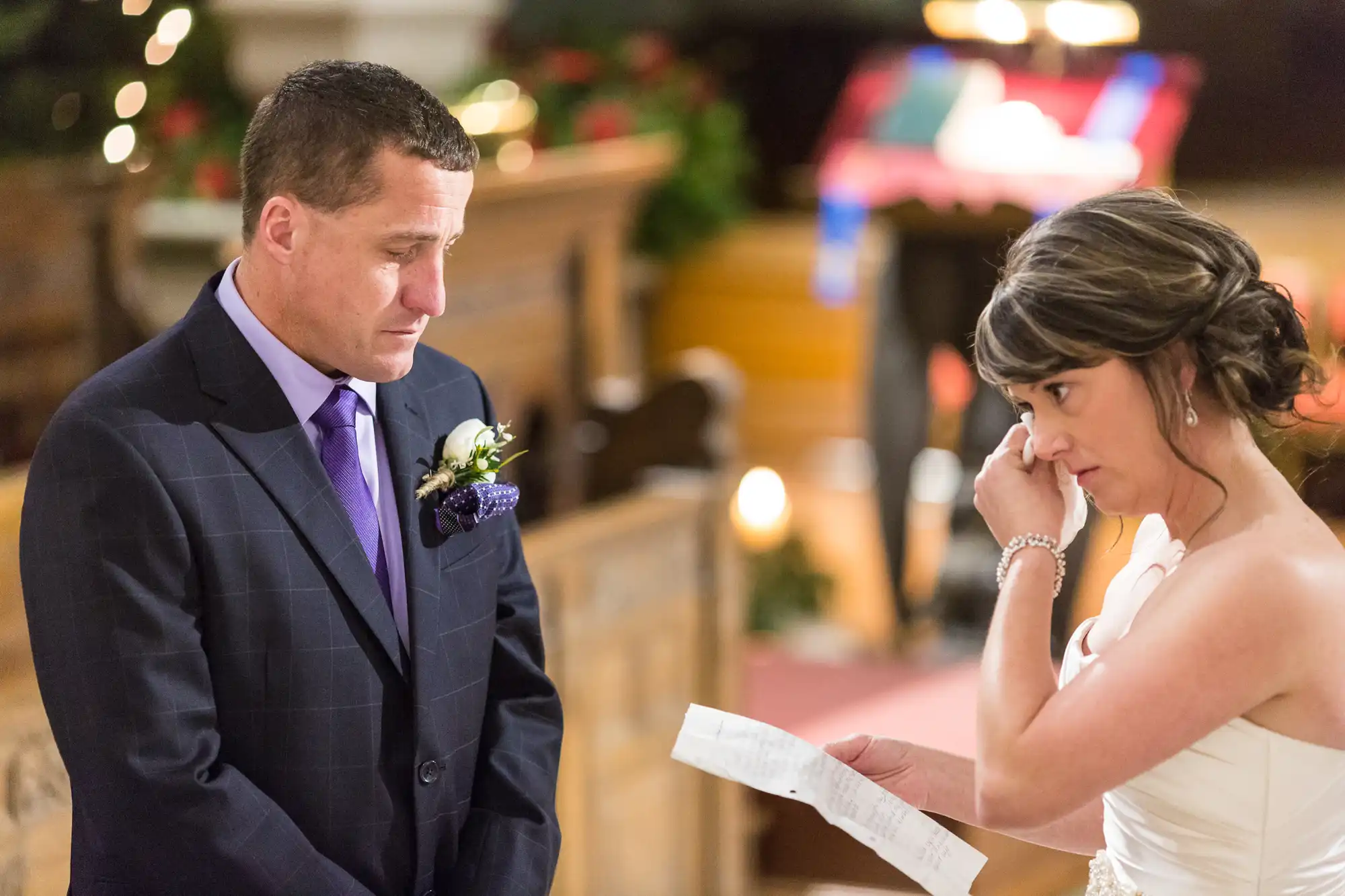 A bride and groom emotionally exchanging vows in a church, the groom looking tearful while the bride reads from a paper.