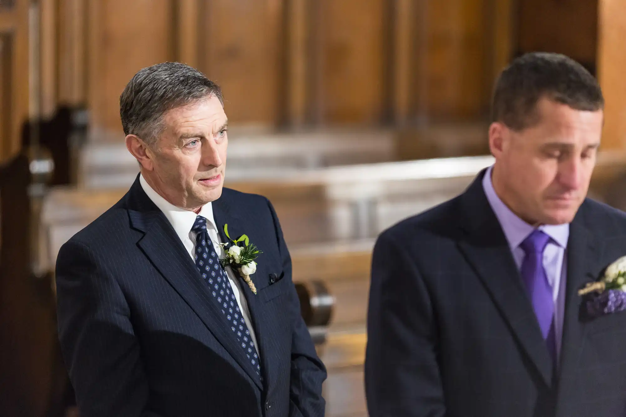Two men in suits with boutonnieres stand solemnly inside a church, one looking intently while the other looks down.