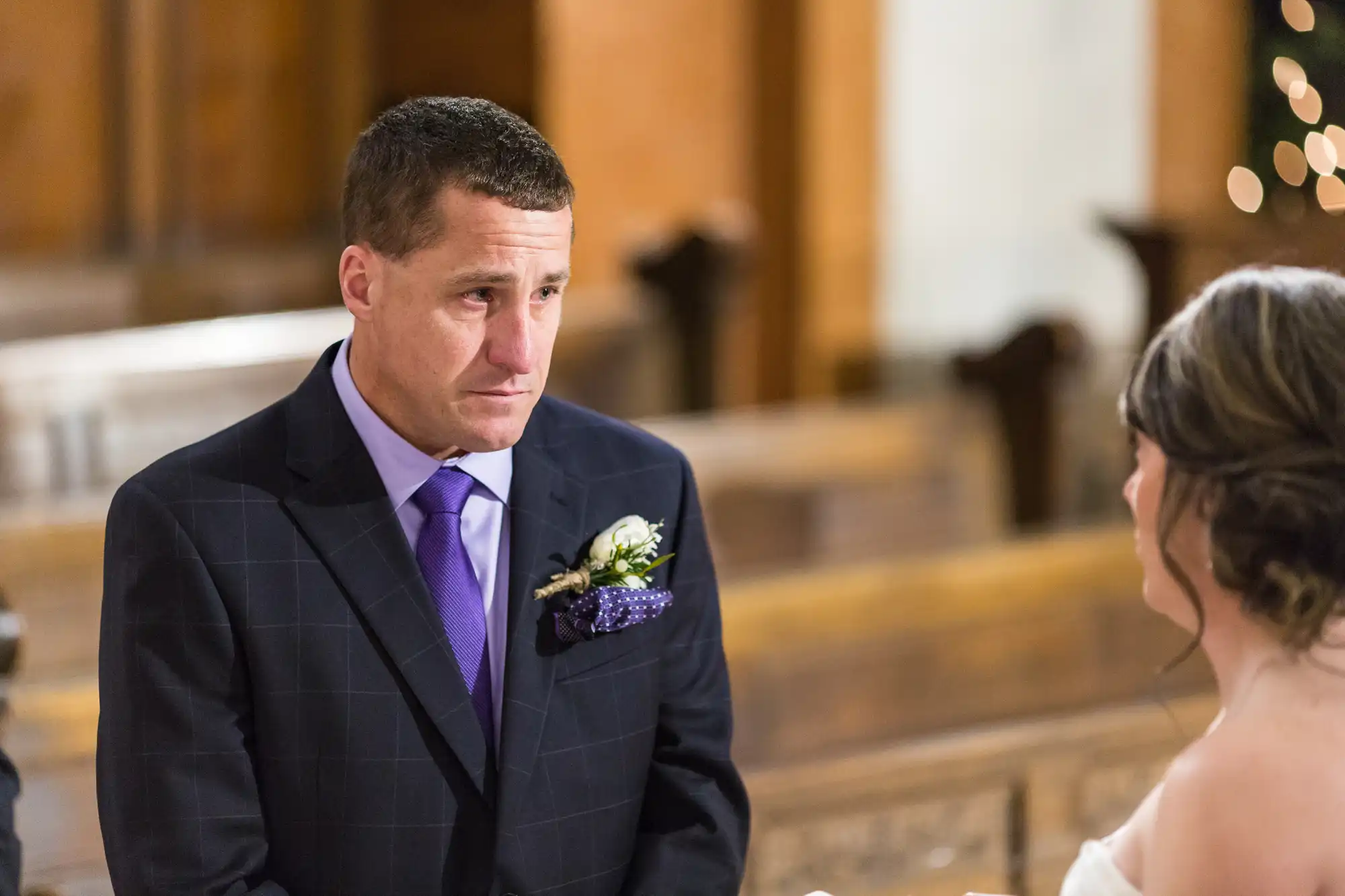 A man in a suit with a boutonniere looks emotionally at a woman in a bridal gown inside a church.