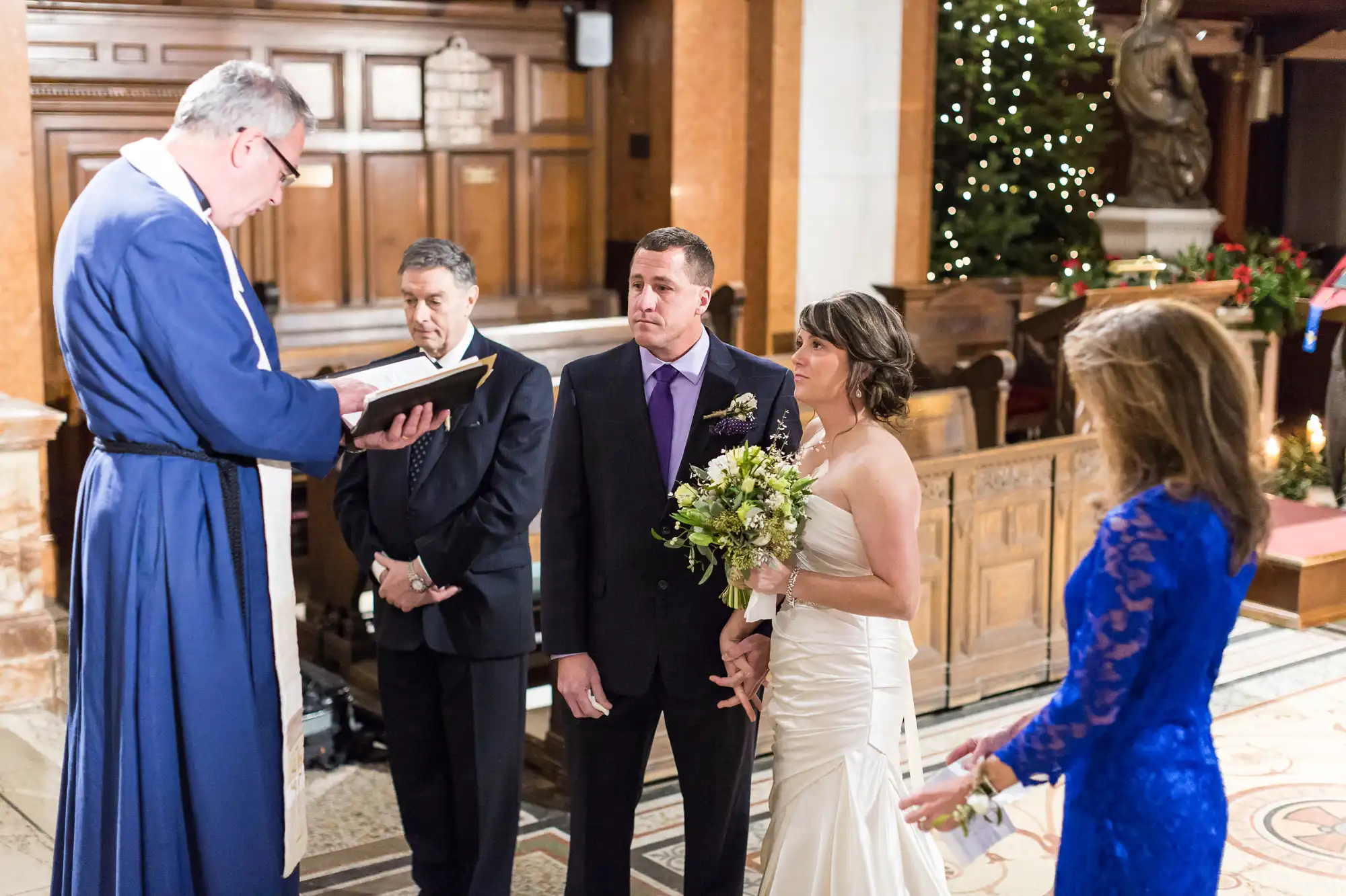 A wedding ceremony inside a church with a priest reading to a bride and groom, and two guests watching.