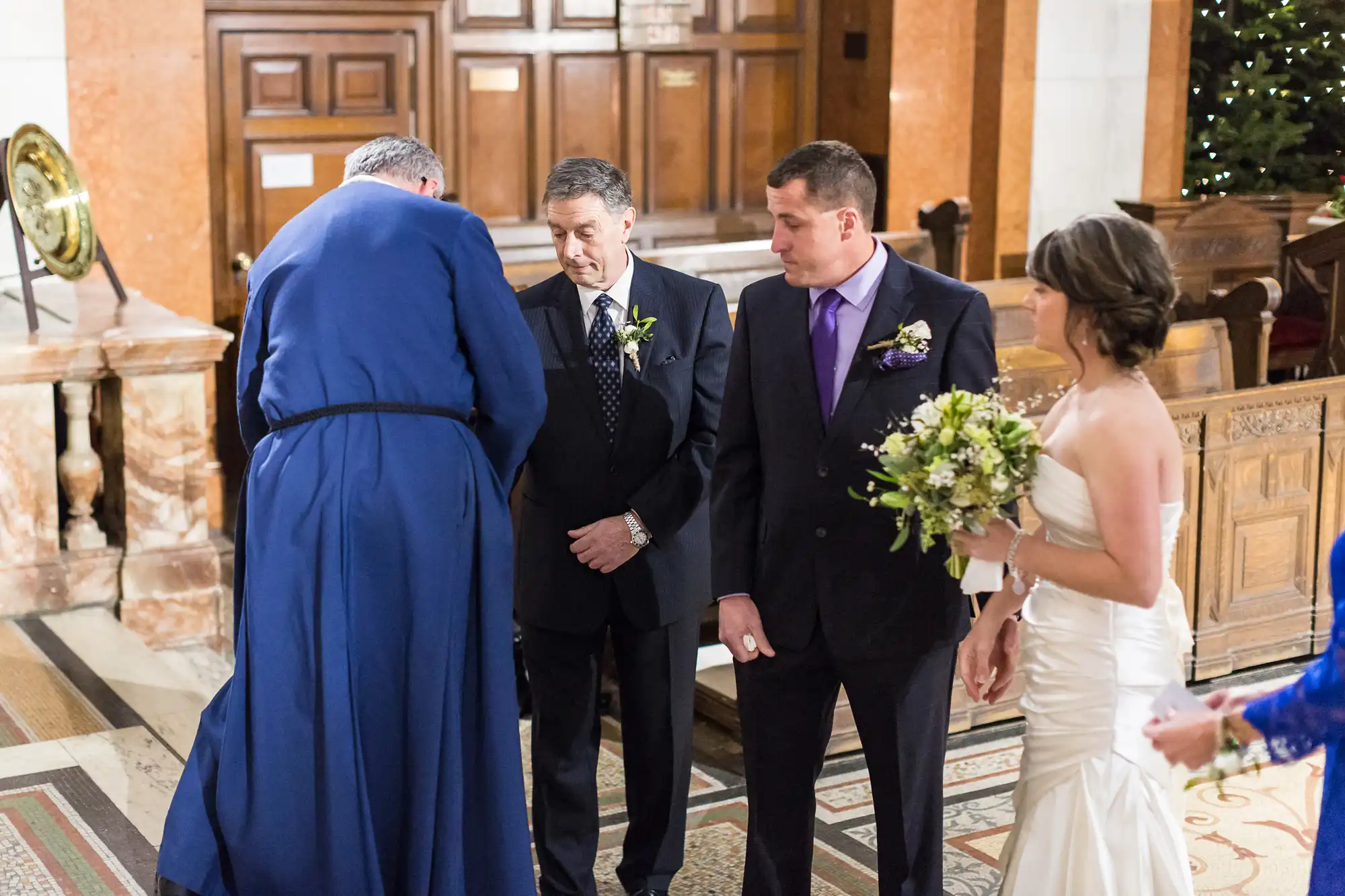 A priest performs a wedding ceremony in a church, standing in front of a groom and best man, with the bride holding a bouquet to the side.