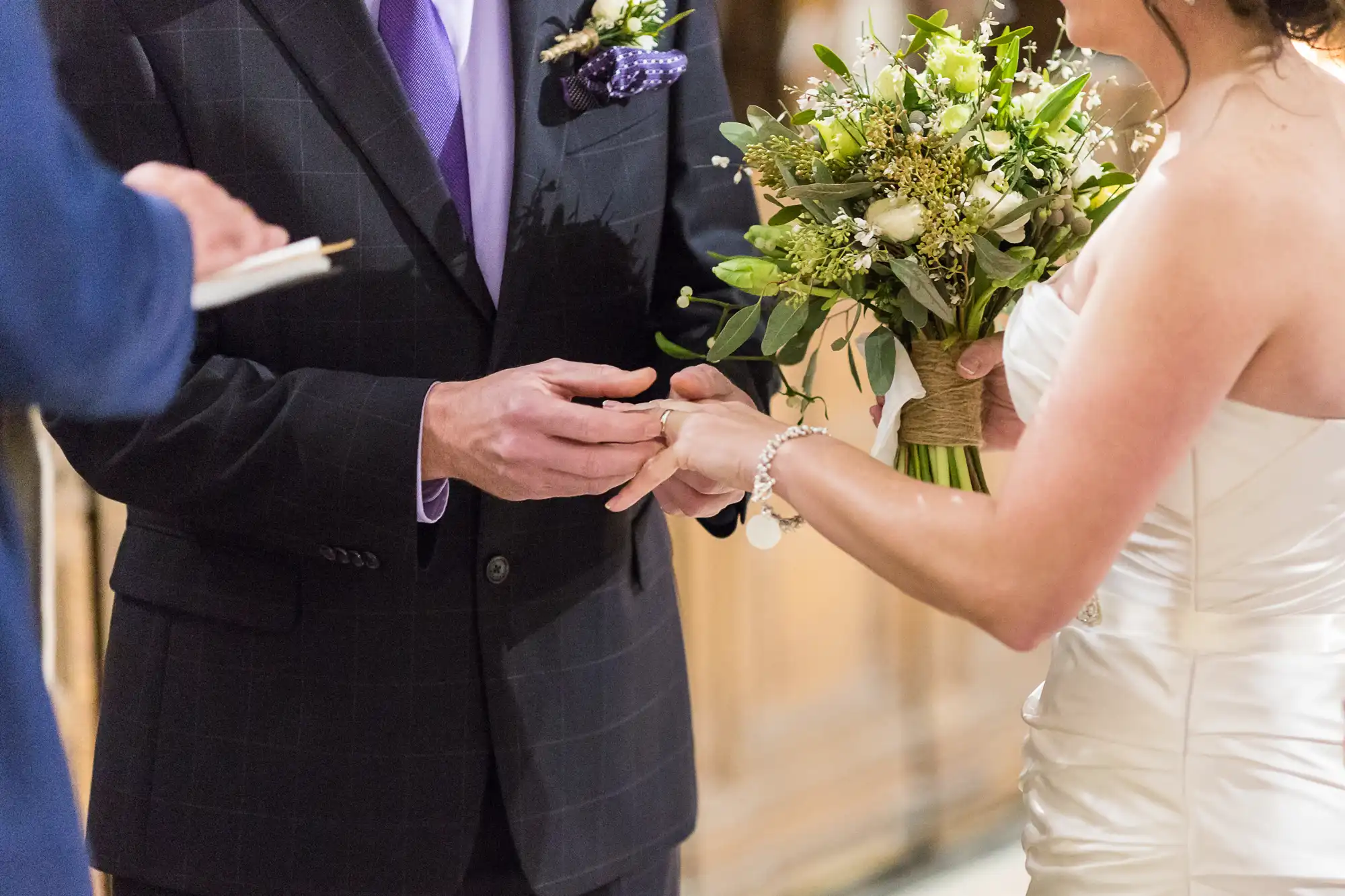 A bride and groom exchange rings during a wedding ceremony, with the groom wearing a purple tie and the bride holding a bouquet.