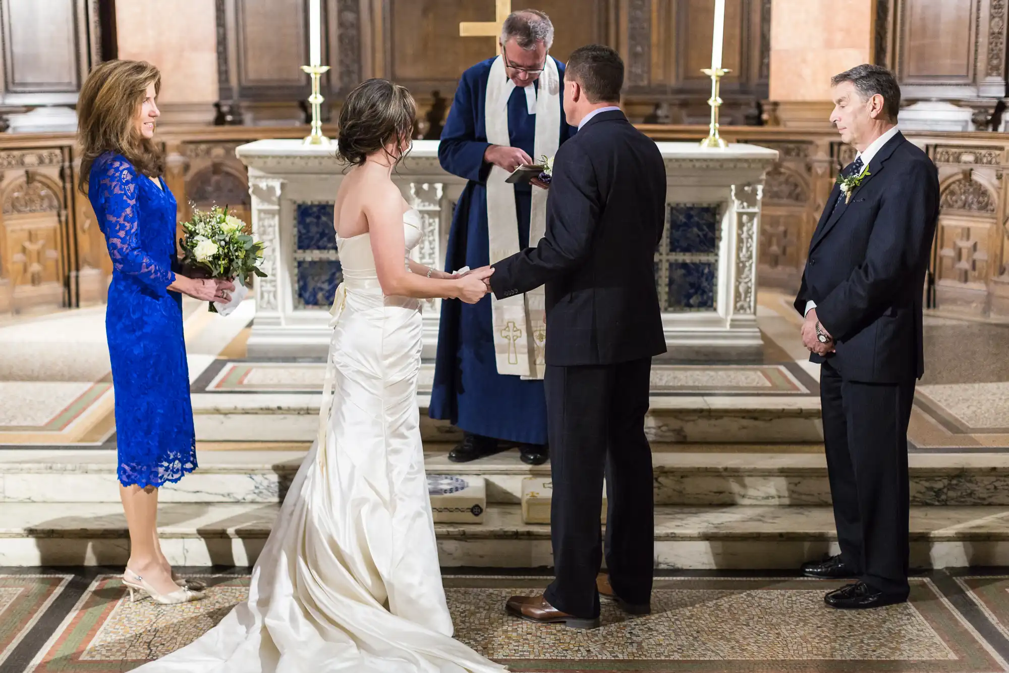 A wedding ceremony in a church with a bride and groom exchanging rings, officiated by a priest, as two witnesses look on.